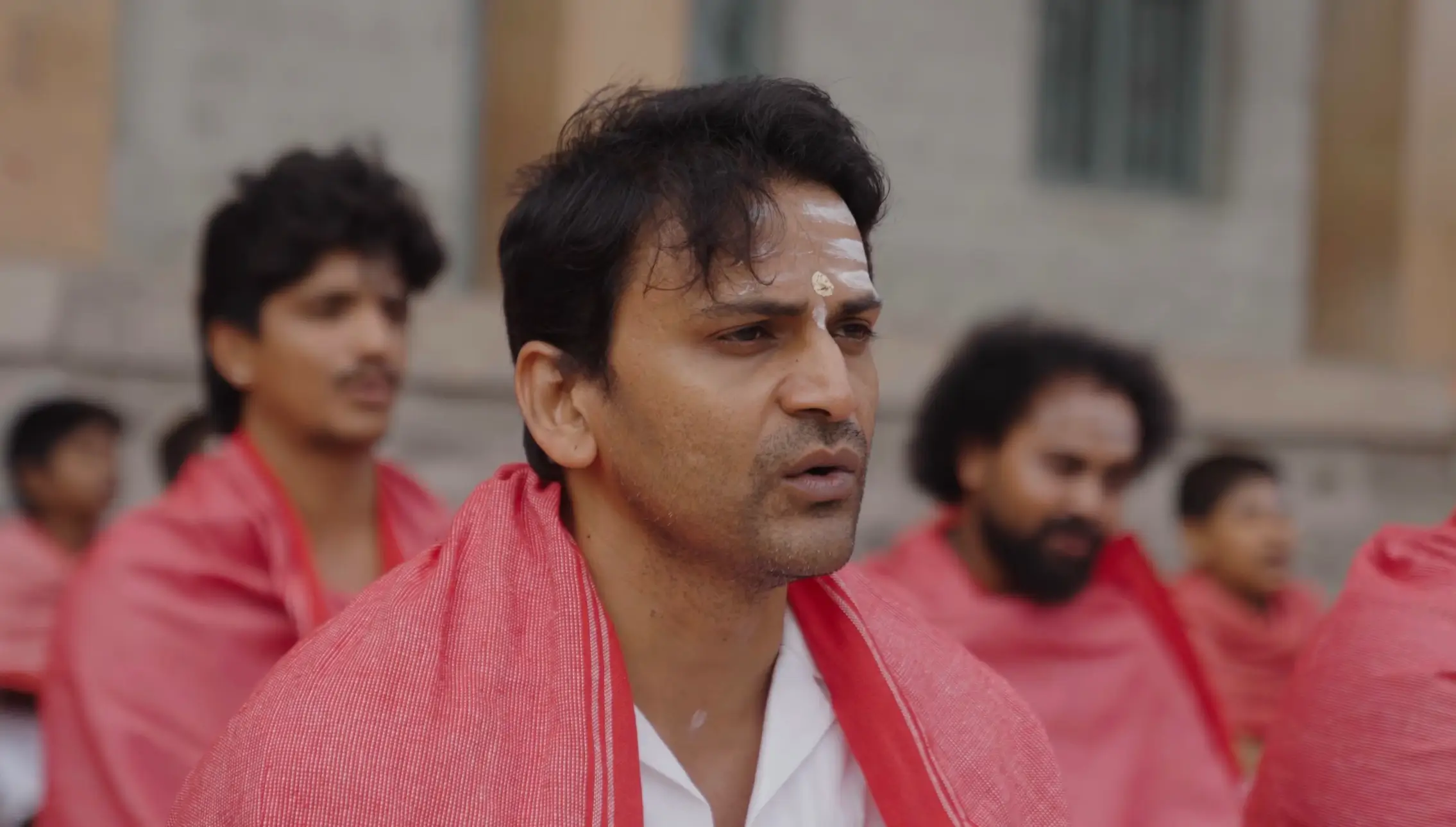 Close-up of a man in a white shirt and red shawl with white forehead markings from the O Belake music video.