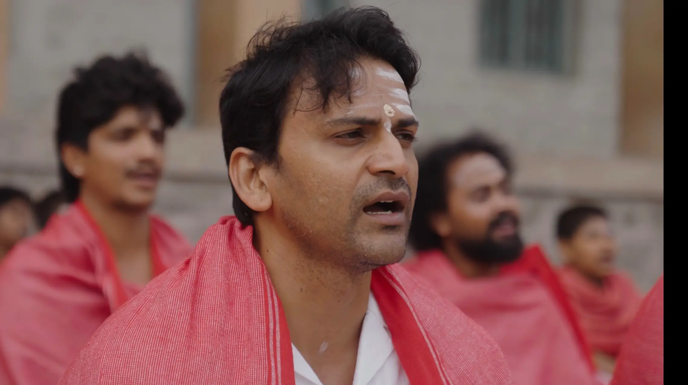 Portrait of a man in a red shawl with white forehead markings from the O Belake music video.