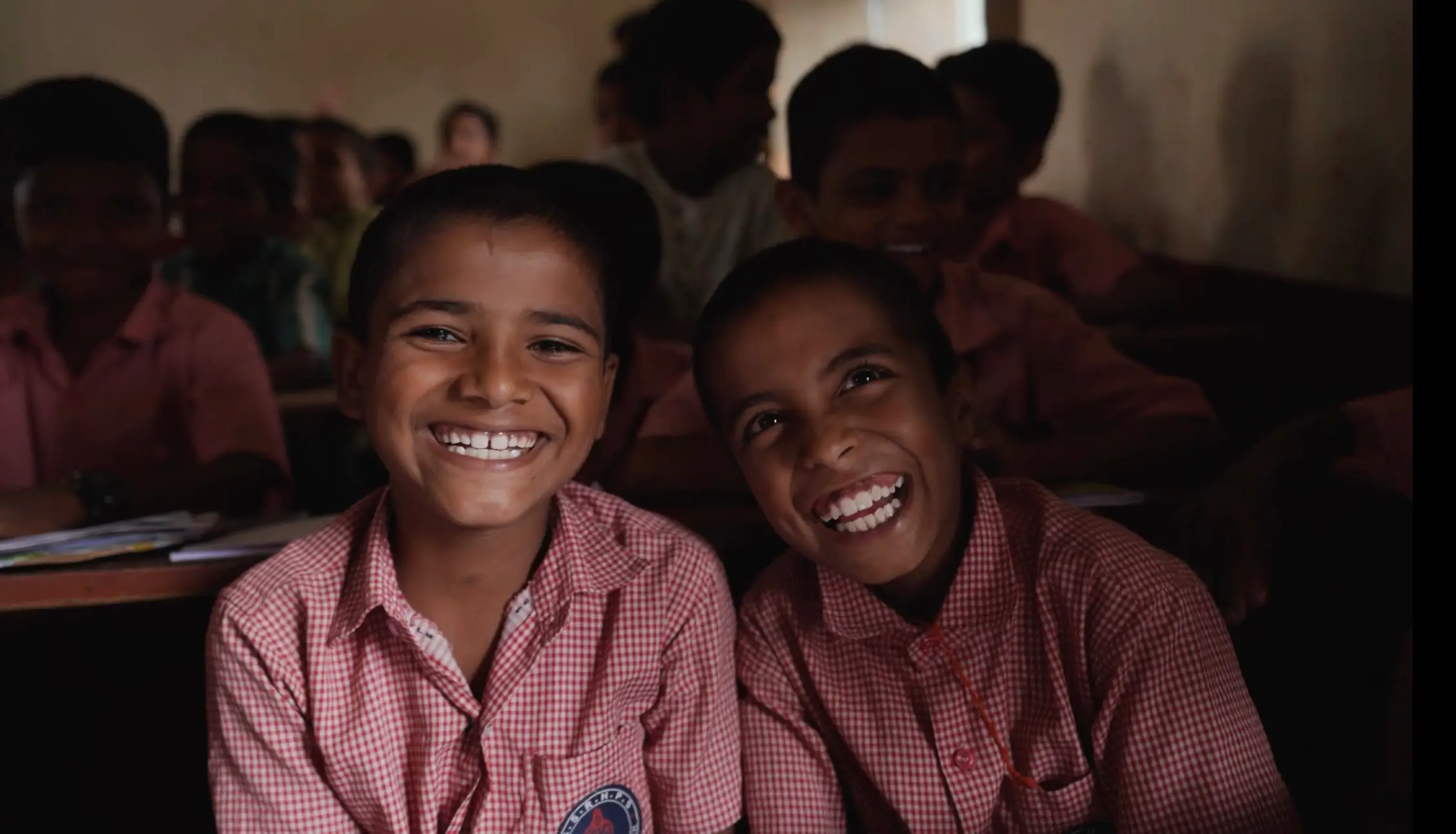 Two smiling schoolchildren in red-and-white checkered uniforms in a classroom from the O Belake music video.