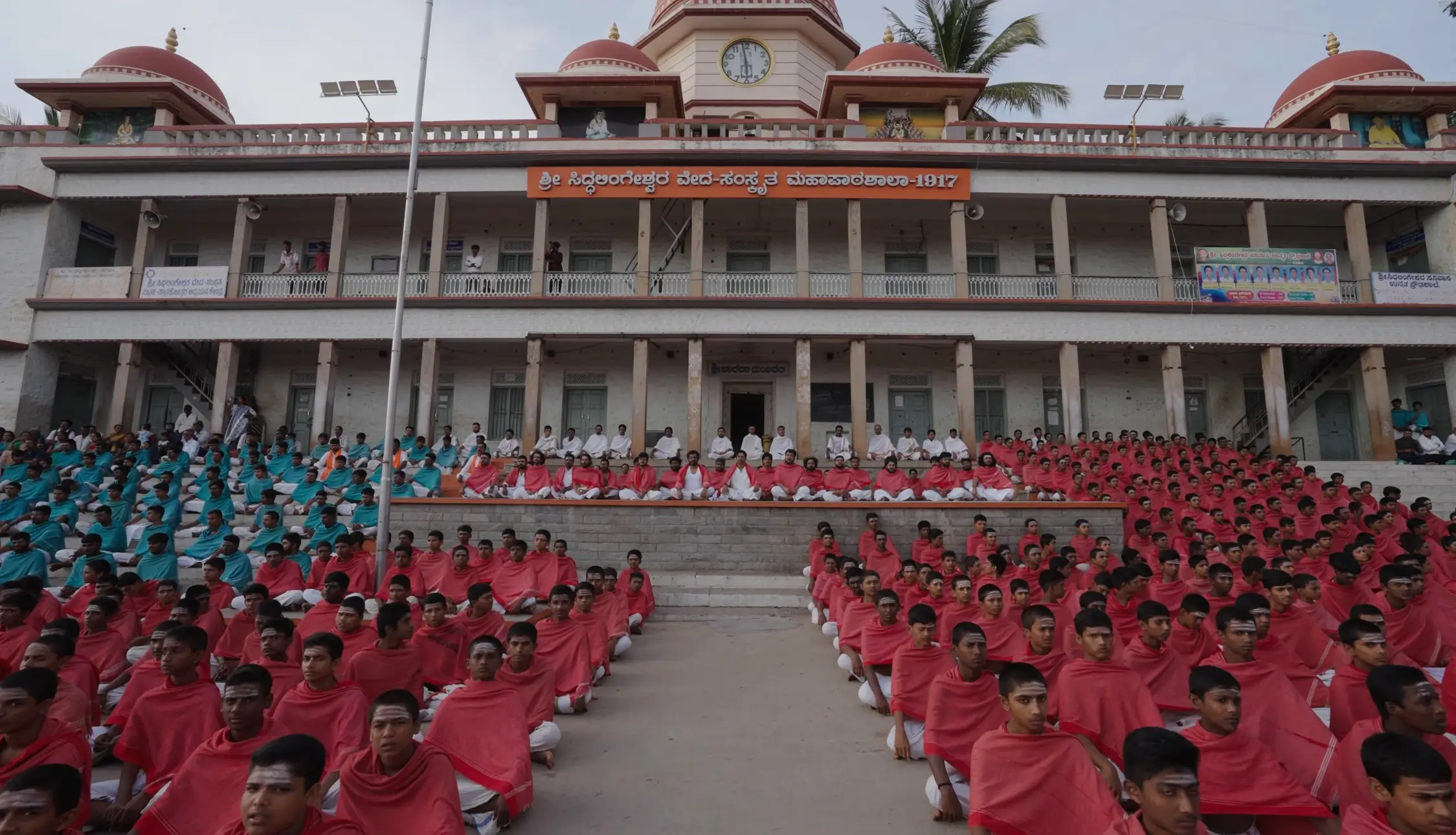 Large gathering in red and teal shawls seated outside a building with domed towers and a clock in the O Belake music video.