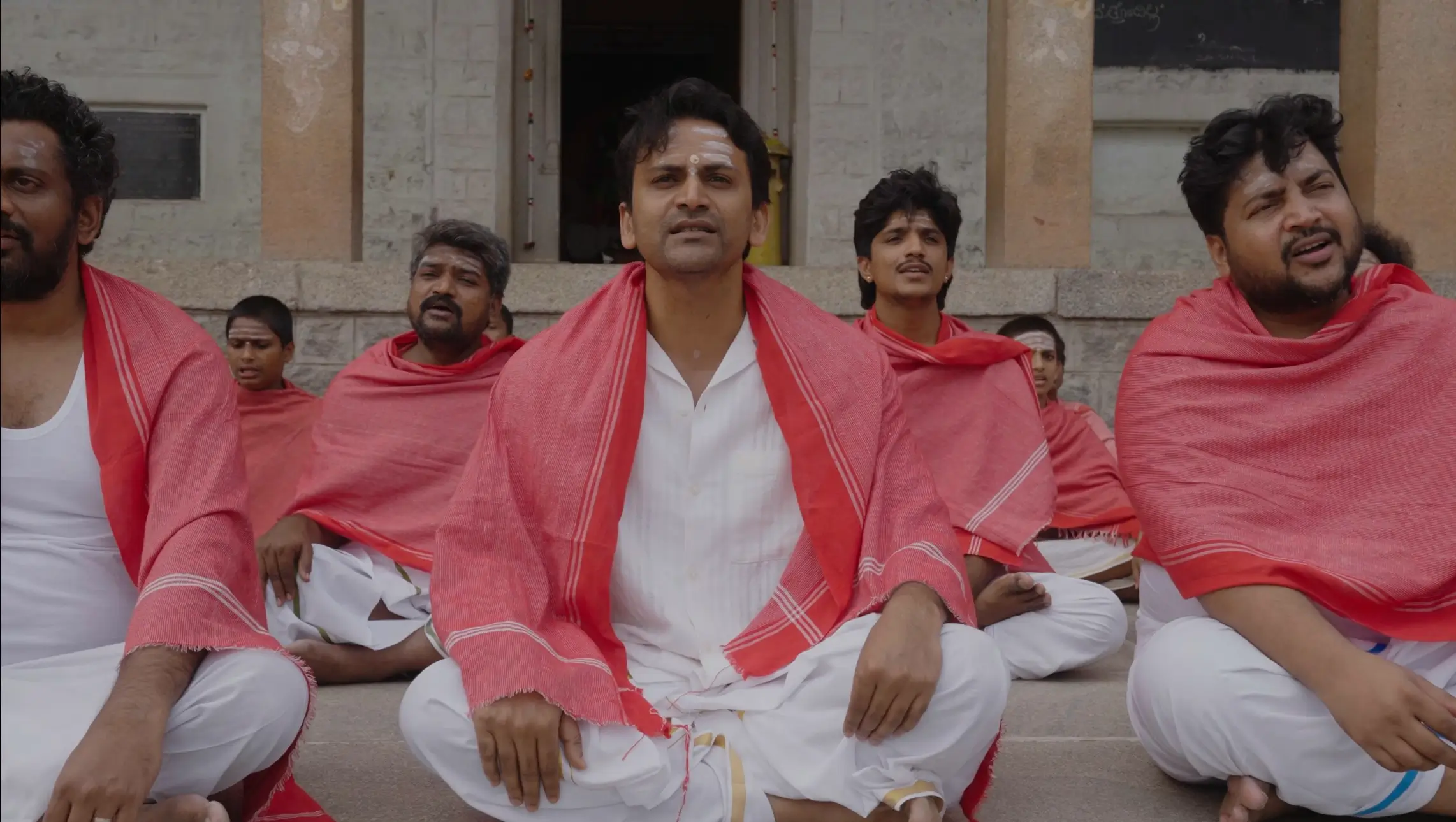 Man in a white shirt and red shawl seated with others on stone steps from the O Belake music video.