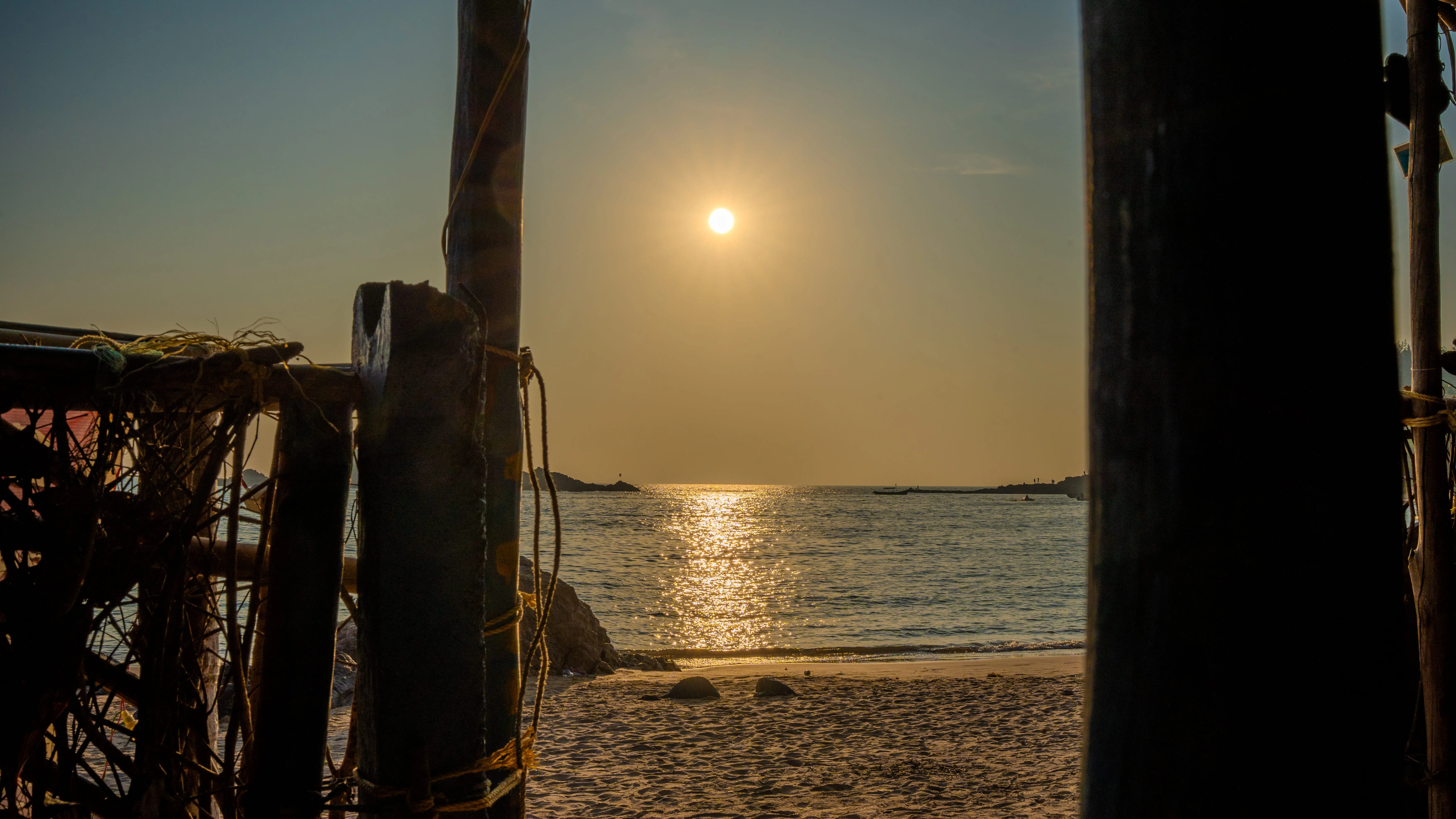 Sunset reflecting across the sea and sand, framed by wooden poles on a Goa beach.