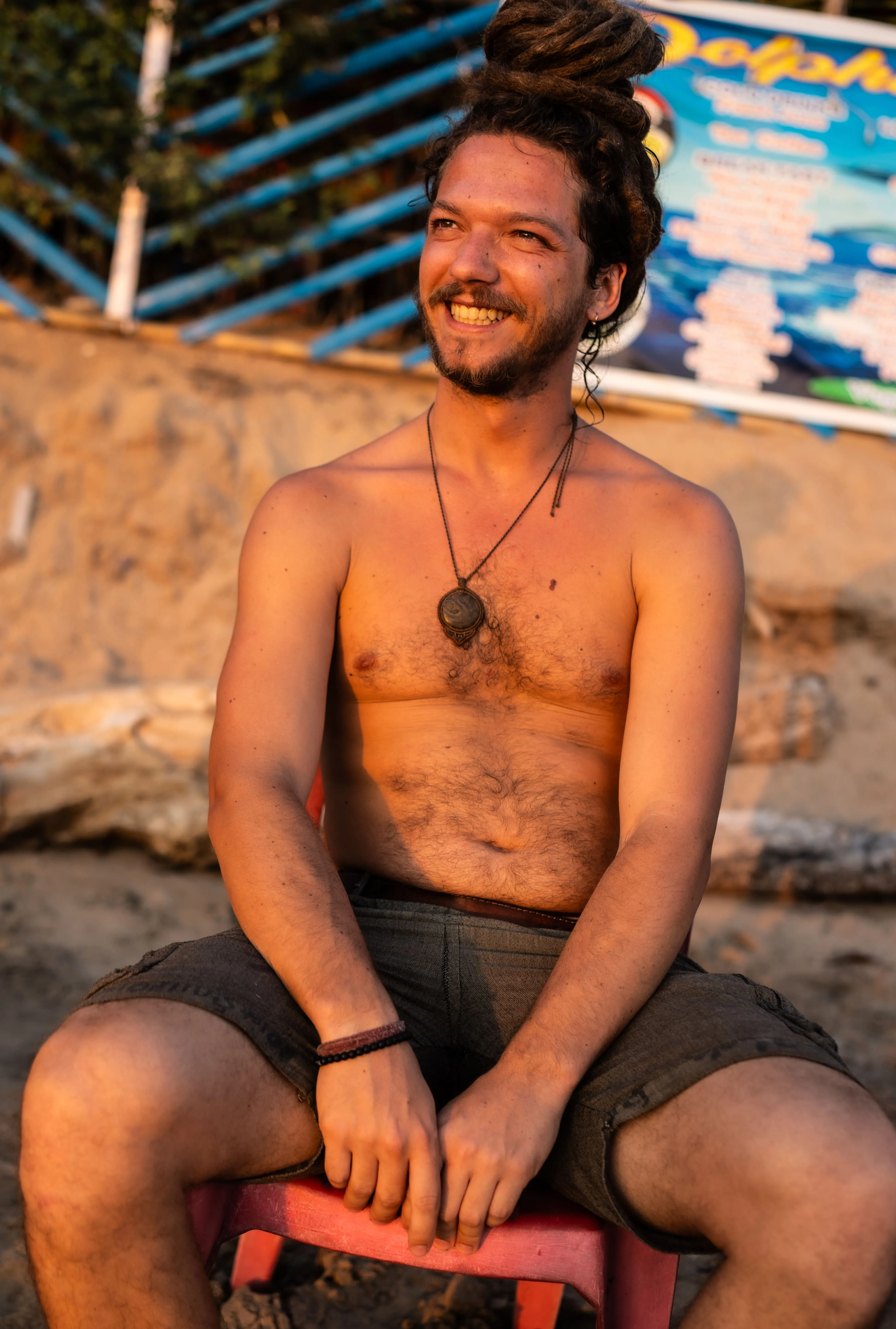 Smiling man with a topknot and pendant necklace seated by a Goa beach.