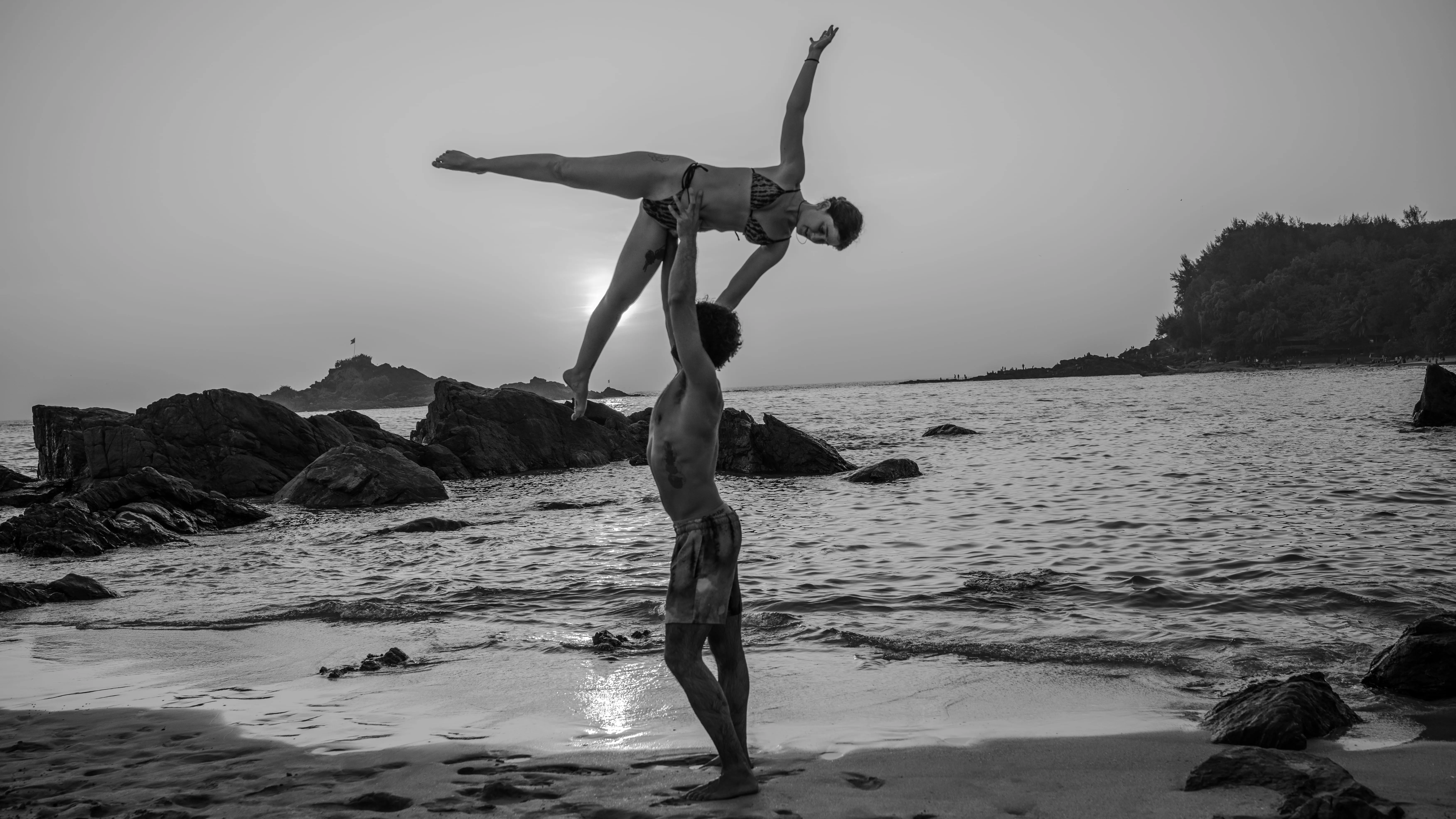 Black-and-white beach photograph of an adult lifting another overhead beside the sea in Goa.
