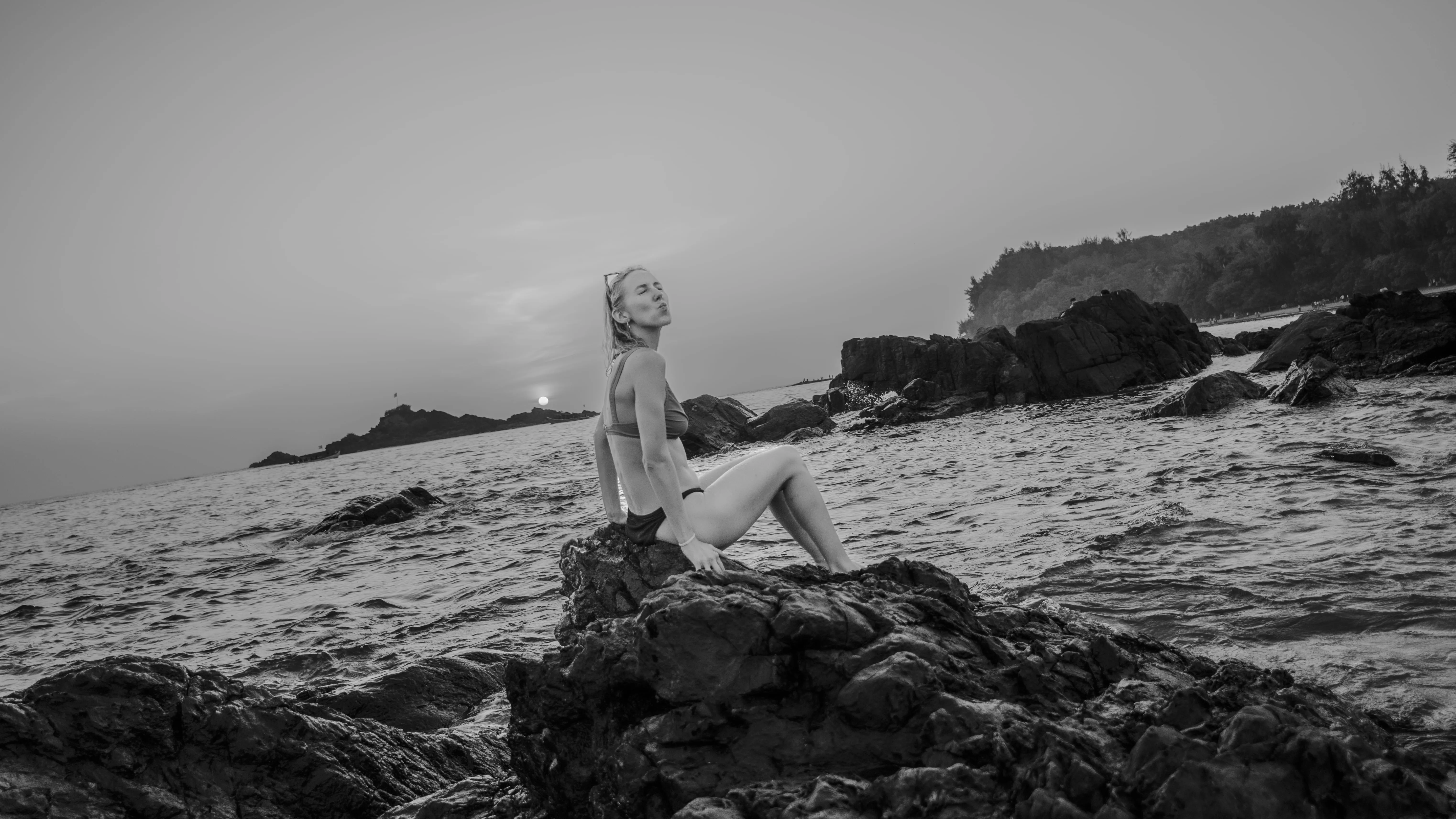 Black-and-white photograph of a woman seated on dark rocks at the water's edge in Goa.