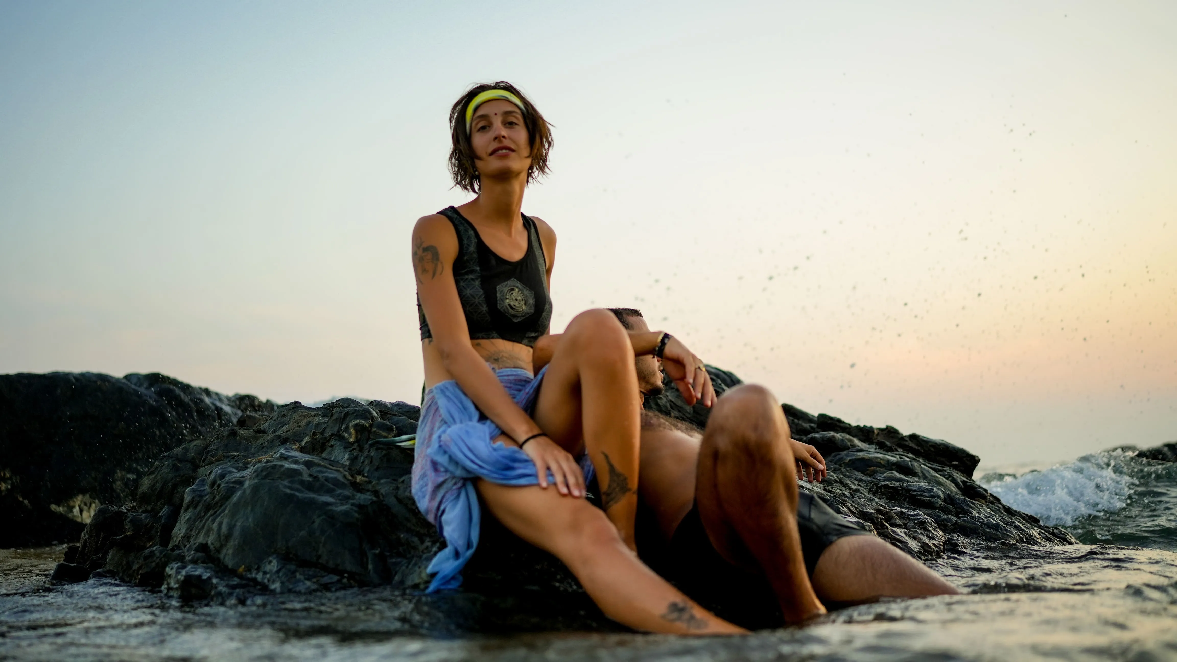 Woman and man seated on dark coastal rocks with sea spray on a Goa beach.