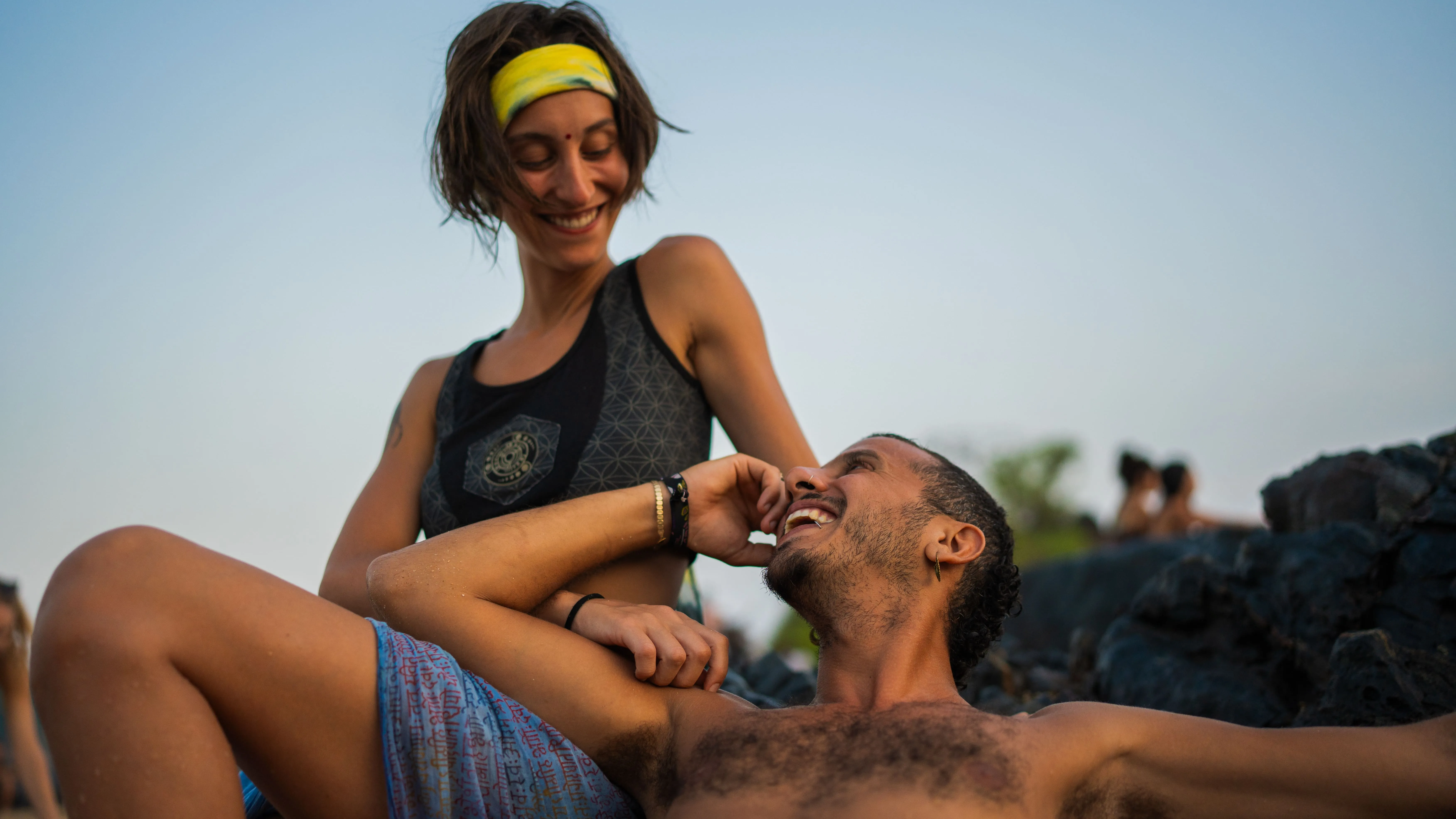 Smiling woman and reclining man on dark coastal rocks at a Goa beach.