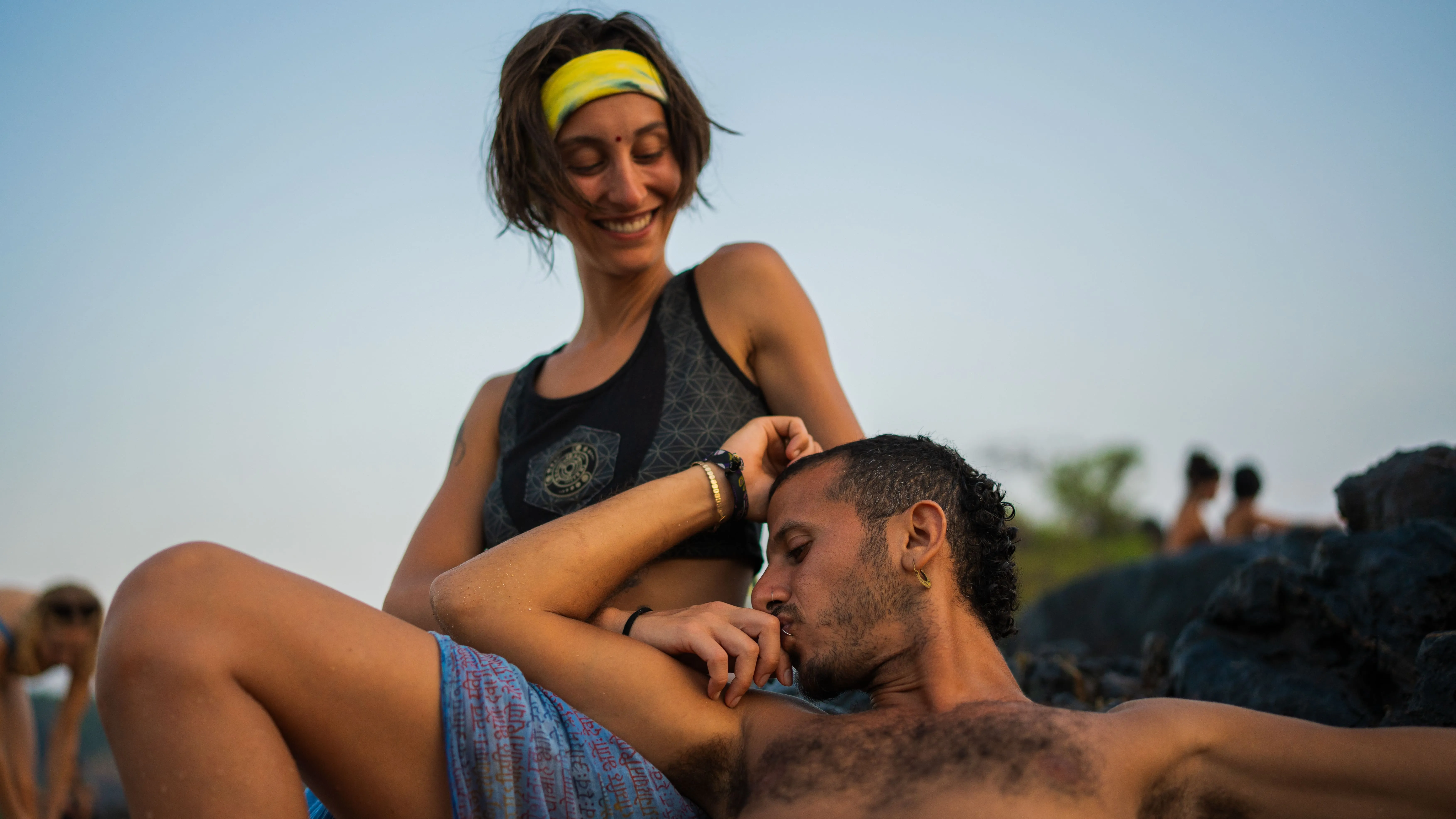 Two adults on dark coastal rocks at a Goa beach, with one resting against the other's leg.