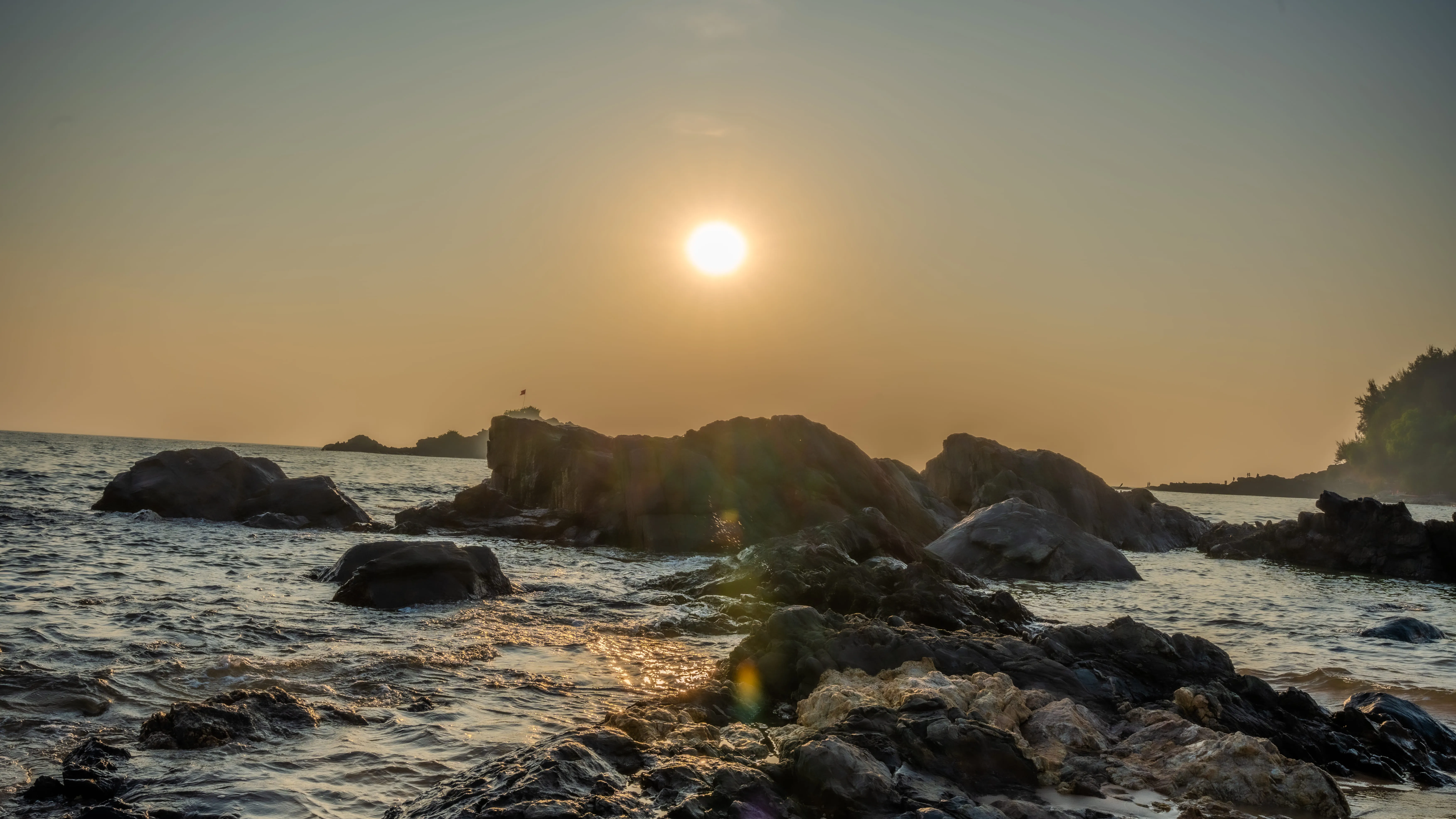 Sunset over waves and dark shoreline rocks on a Goa beach.