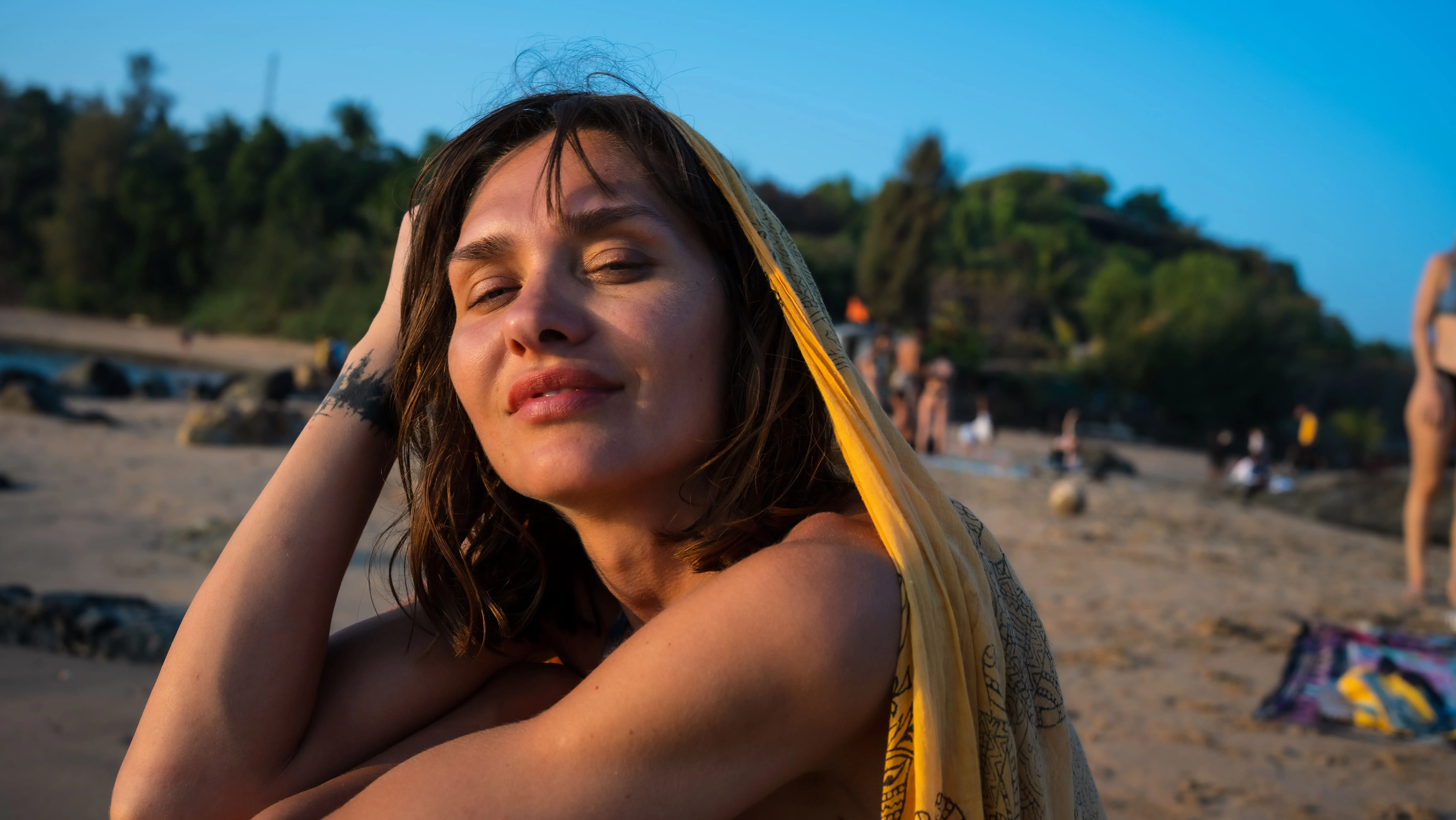 Woman in a yellow patterned scarf with one hand in her hair on a Goa beach.