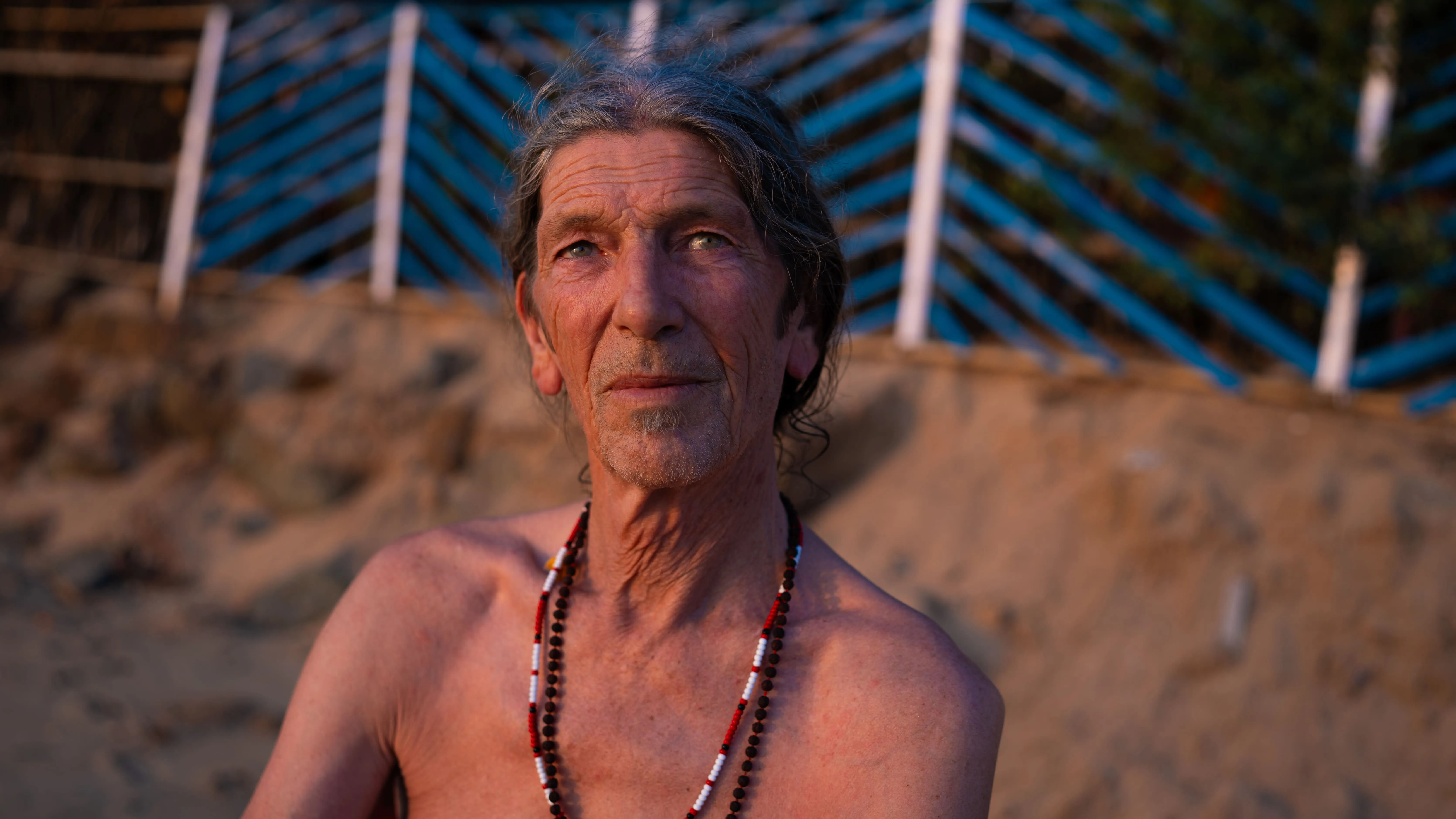 Sunlit portrait of an older man wearing layered bead necklaces on a Goa beach.