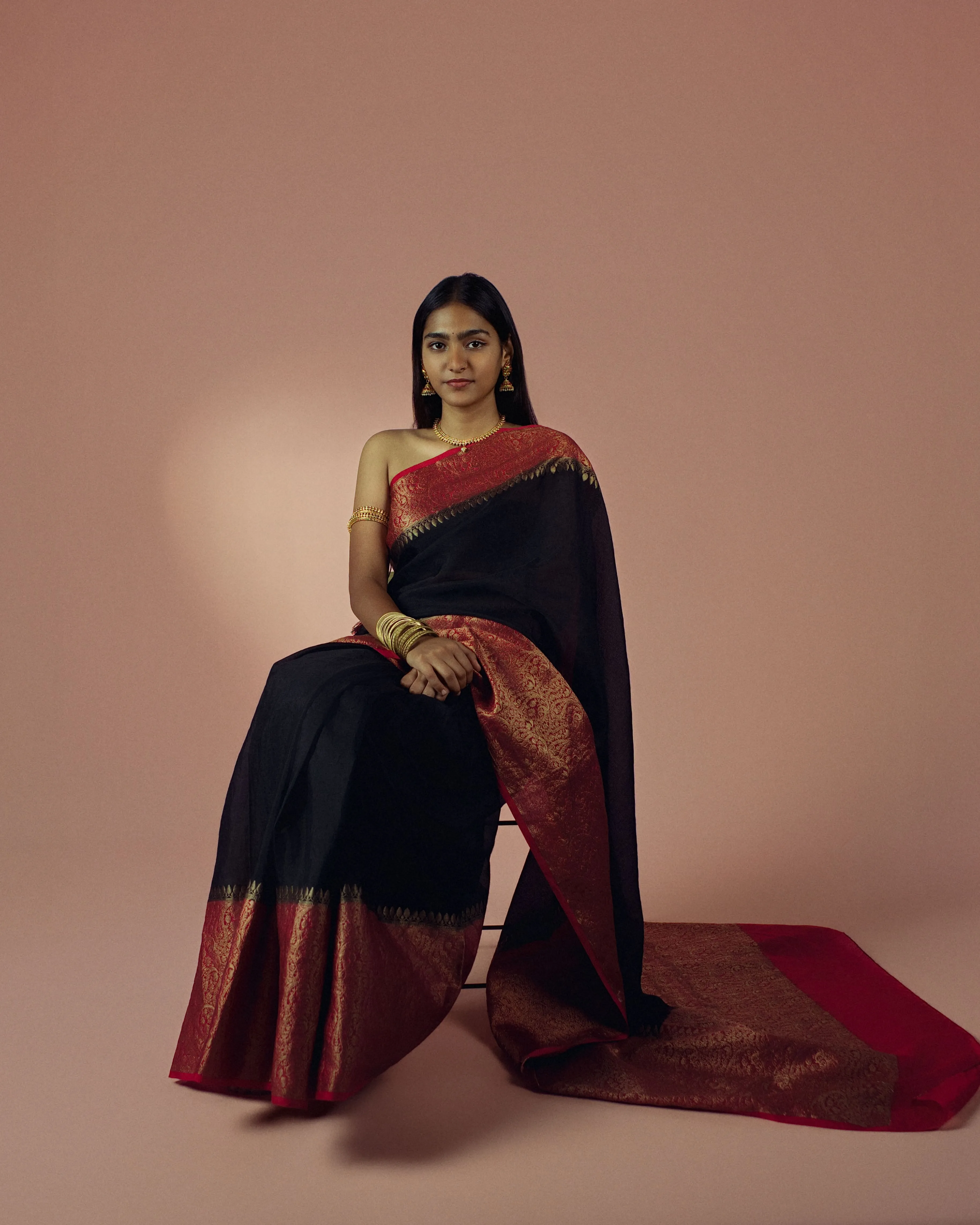 Full-length portrait of a seated woman in a black sari with a red and gold border.