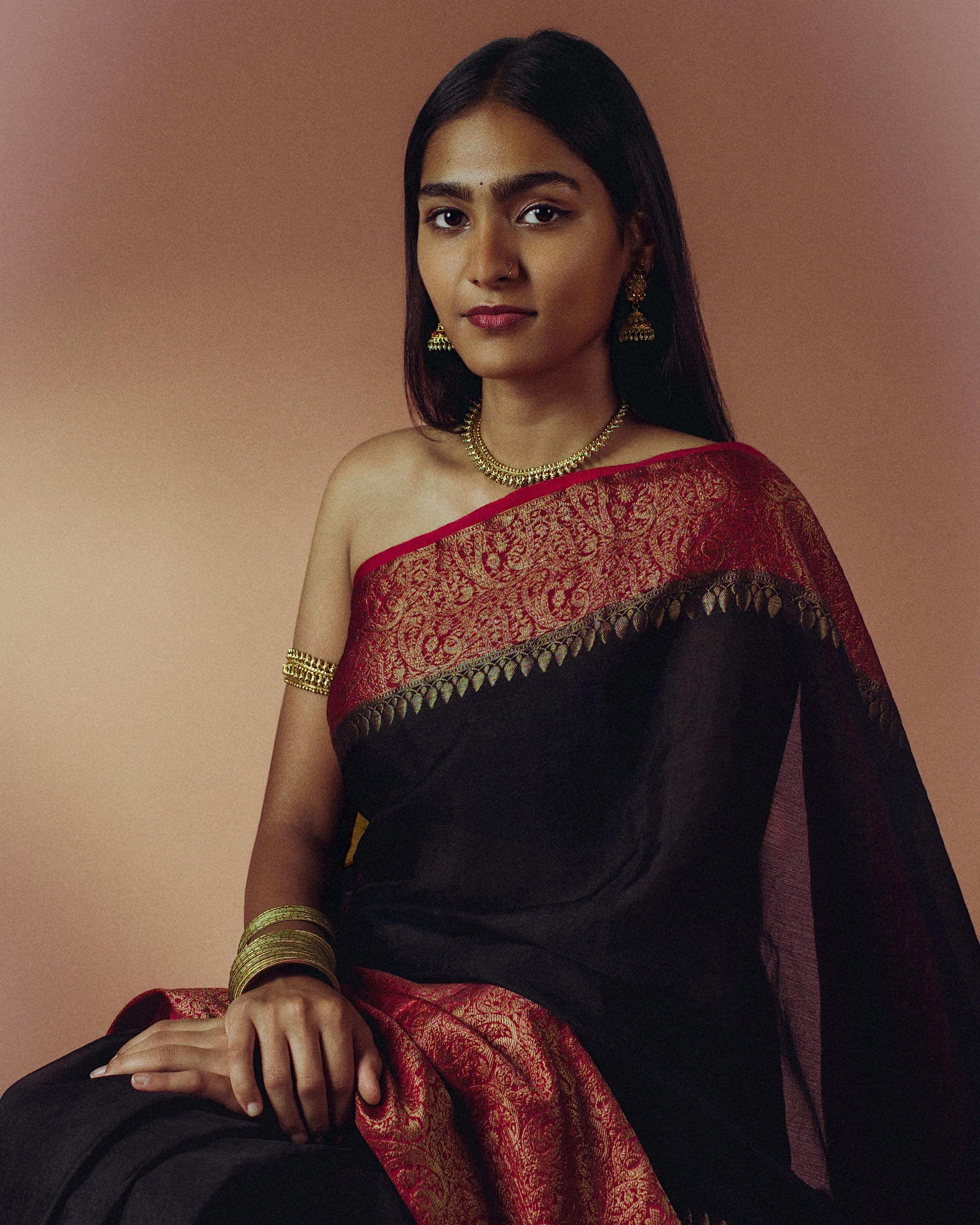 Seated studio portrait of a woman in a black sari with a red and gold border.