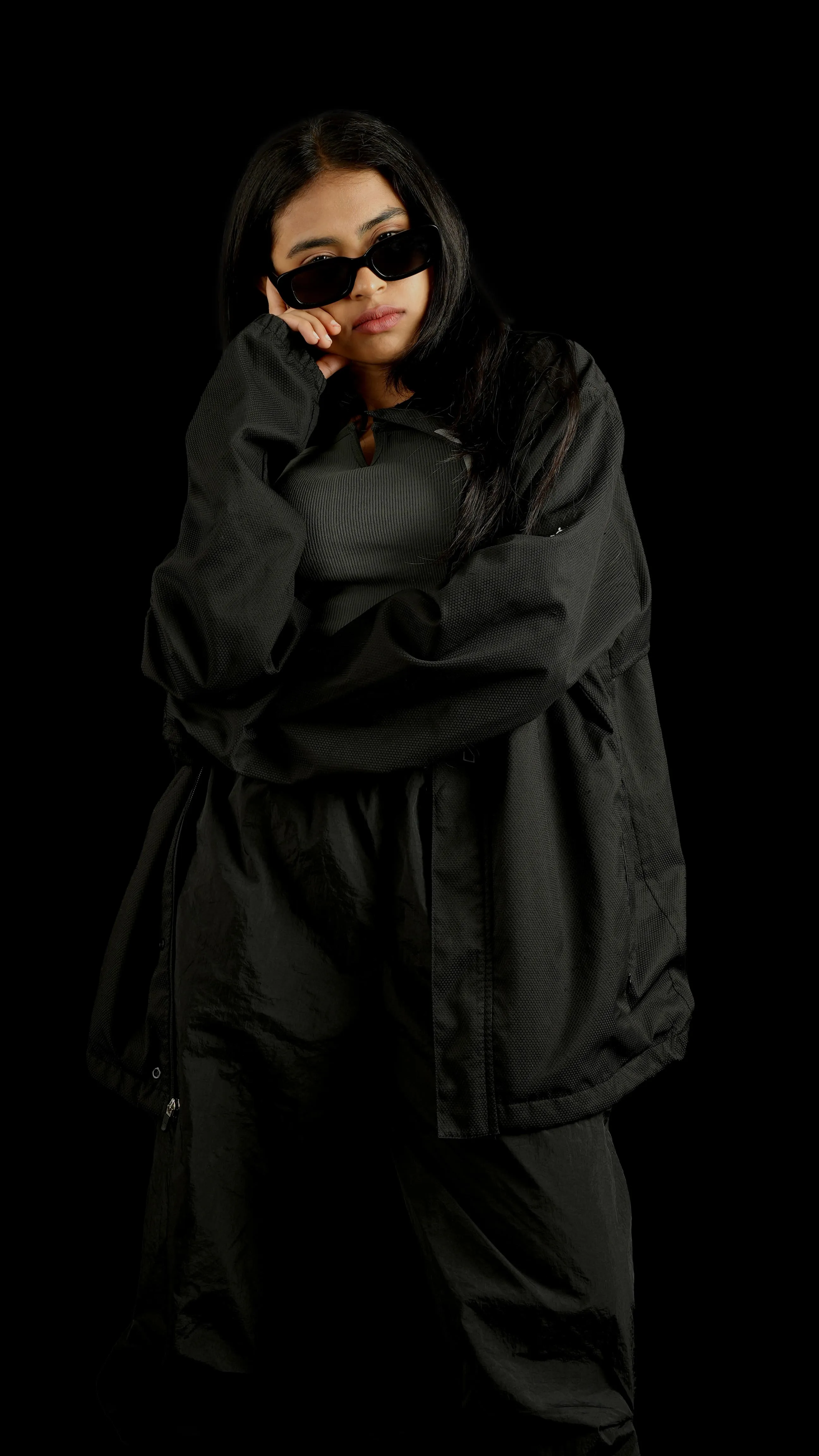 Woman in black sunglasses and a black jacket with arms crossed in a studio portrait.