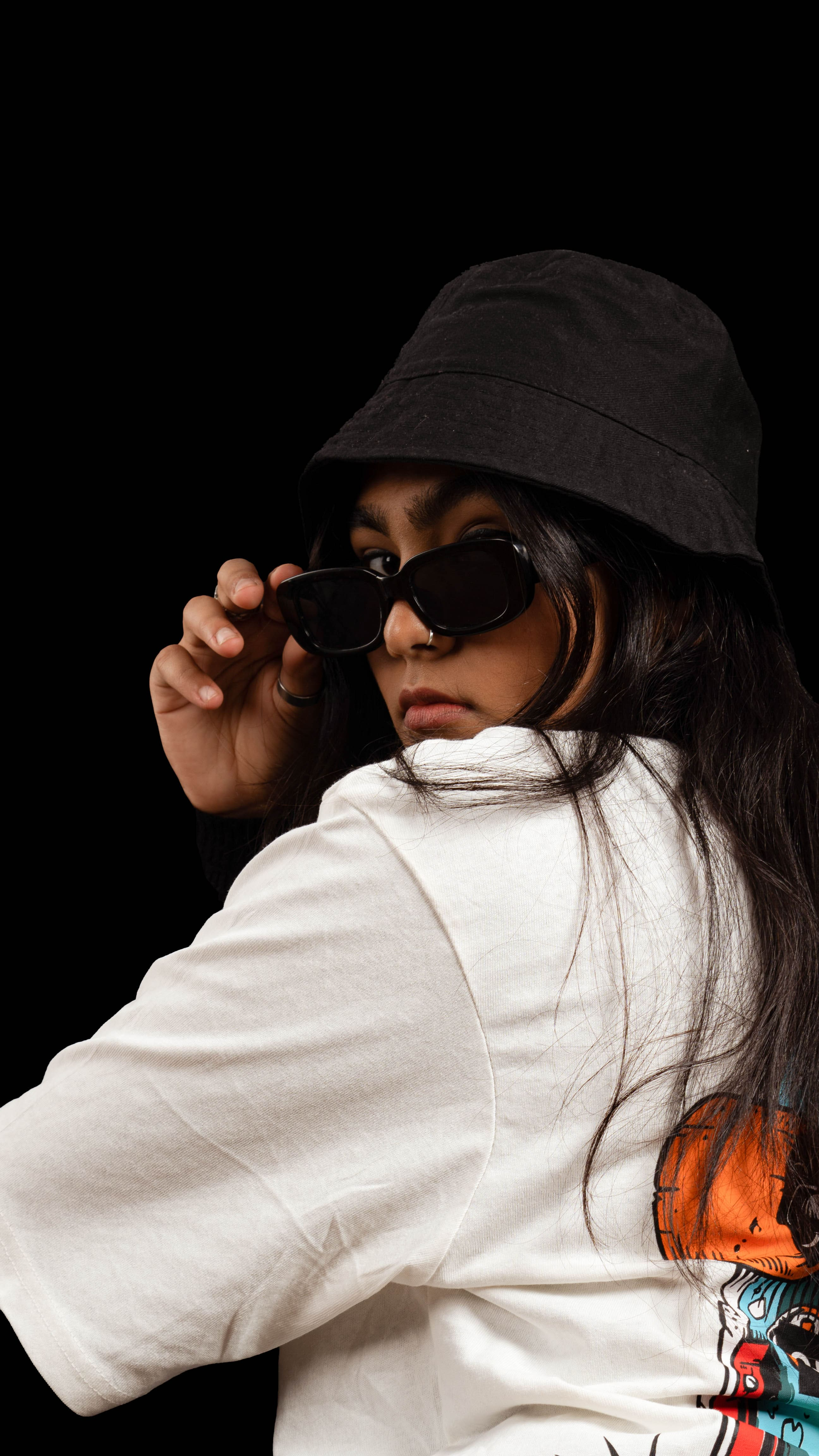 Woman in a black bucket hat leaning forward to adjust her sunglasses over a white graphic T-shirt.