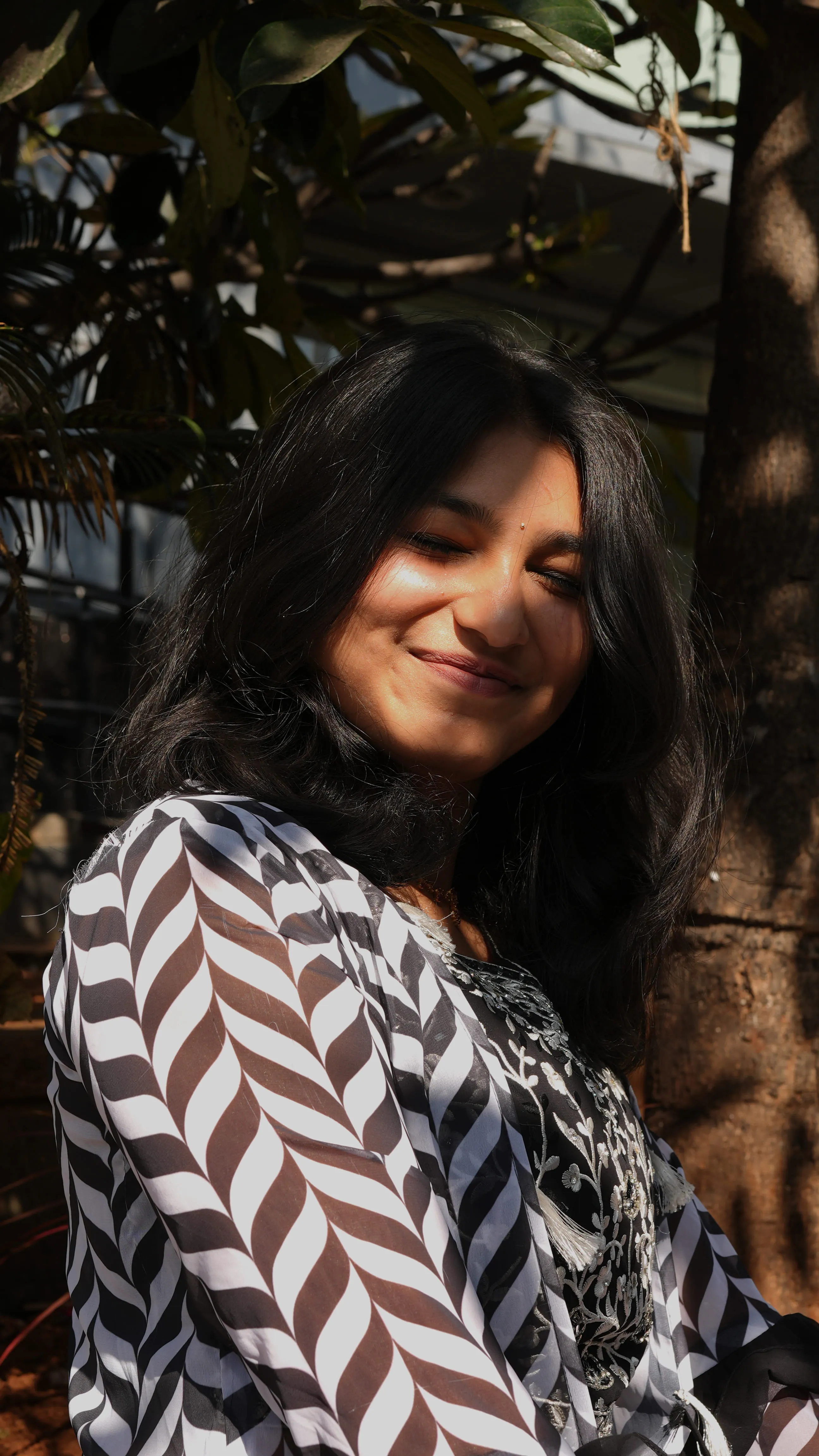 Sunlit outdoor portrait of a smiling woman in a black-and-white patterned top.