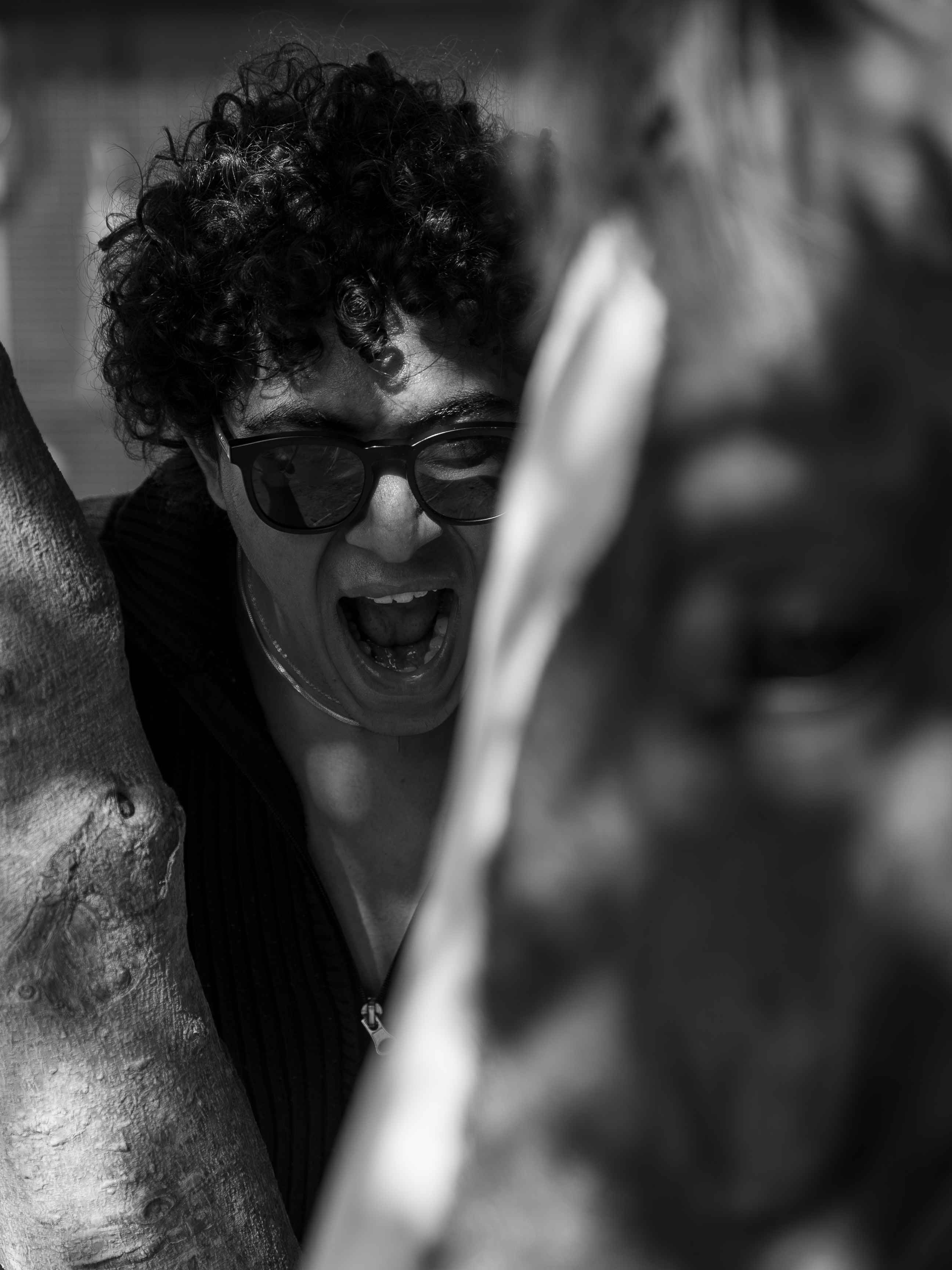 Black-and-white candid portrait of a person in glasses framed by a tree trunk.