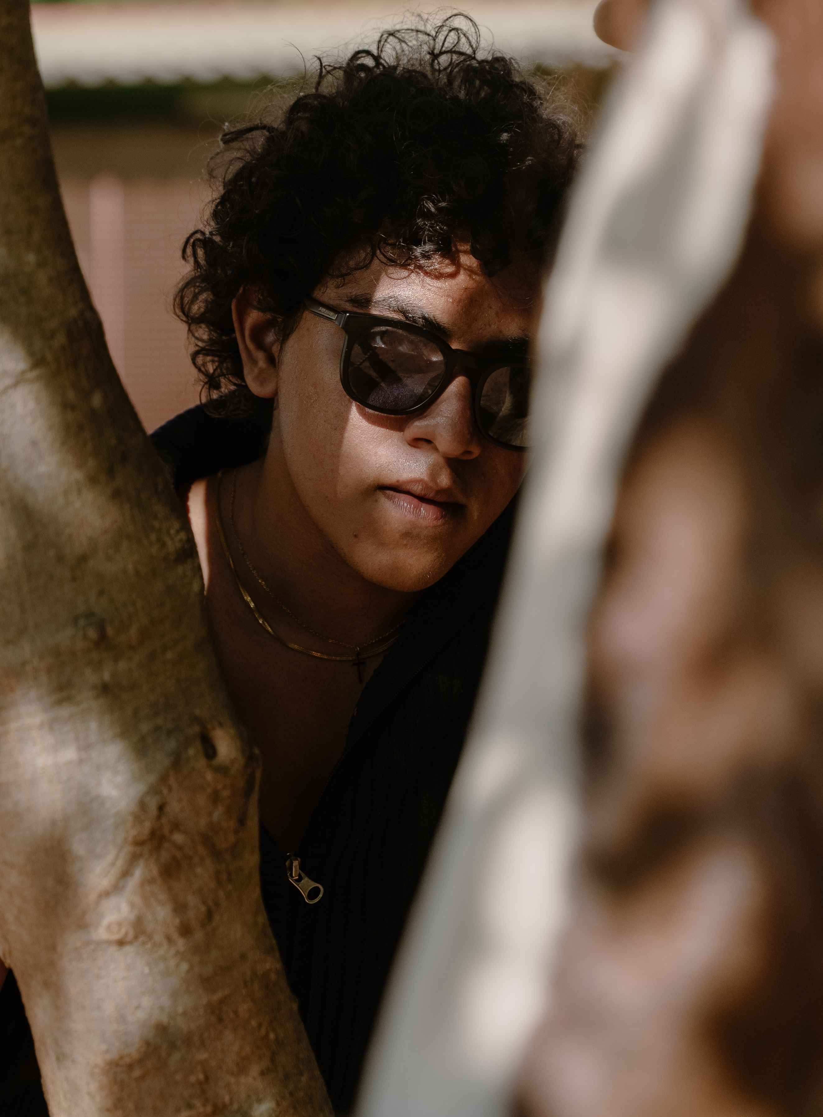 Outdoor portrait of a person in dark glasses framed by a tree trunk.