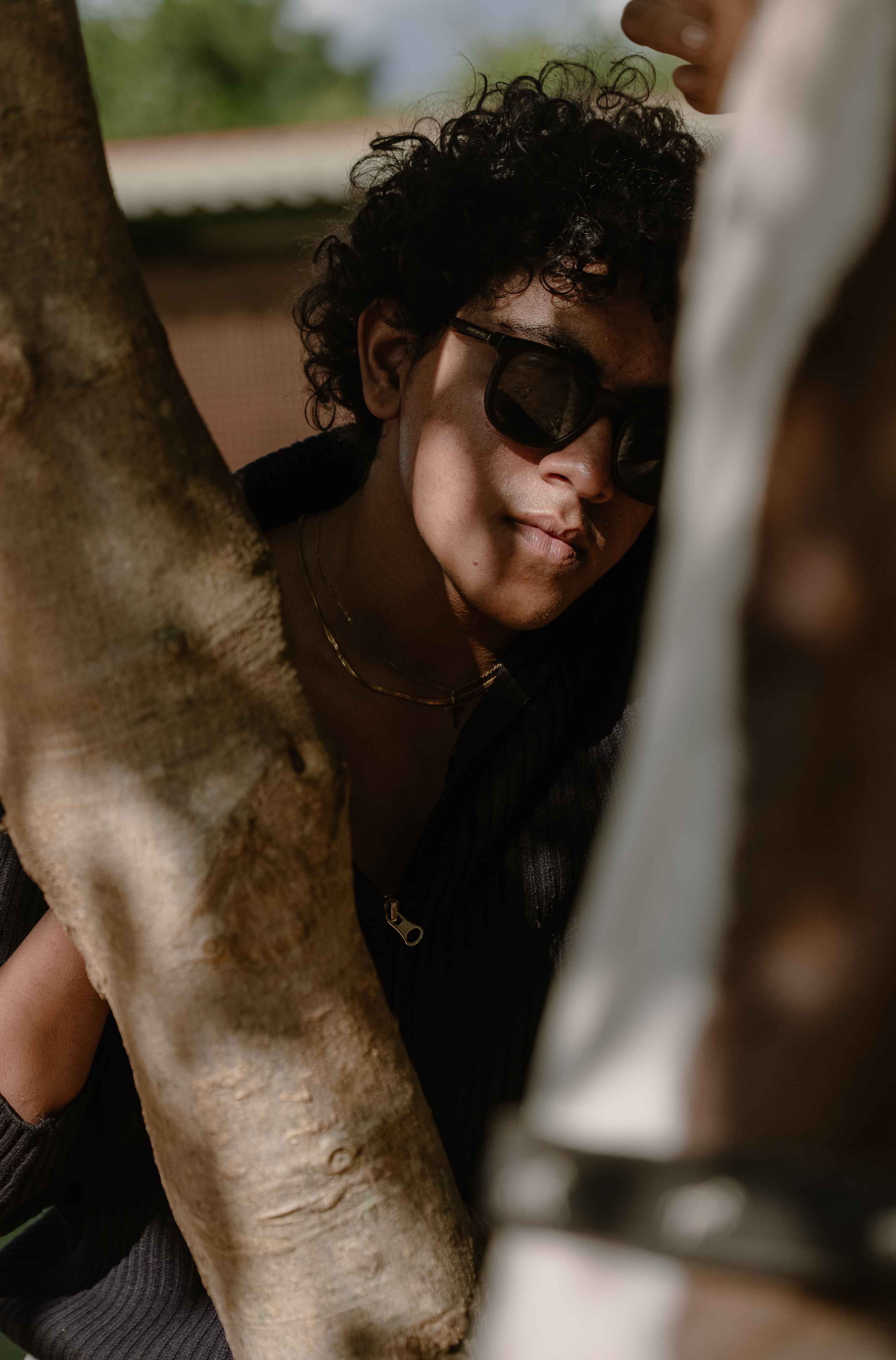 Outdoor portrait of a person in dark glasses beside a tree trunk.