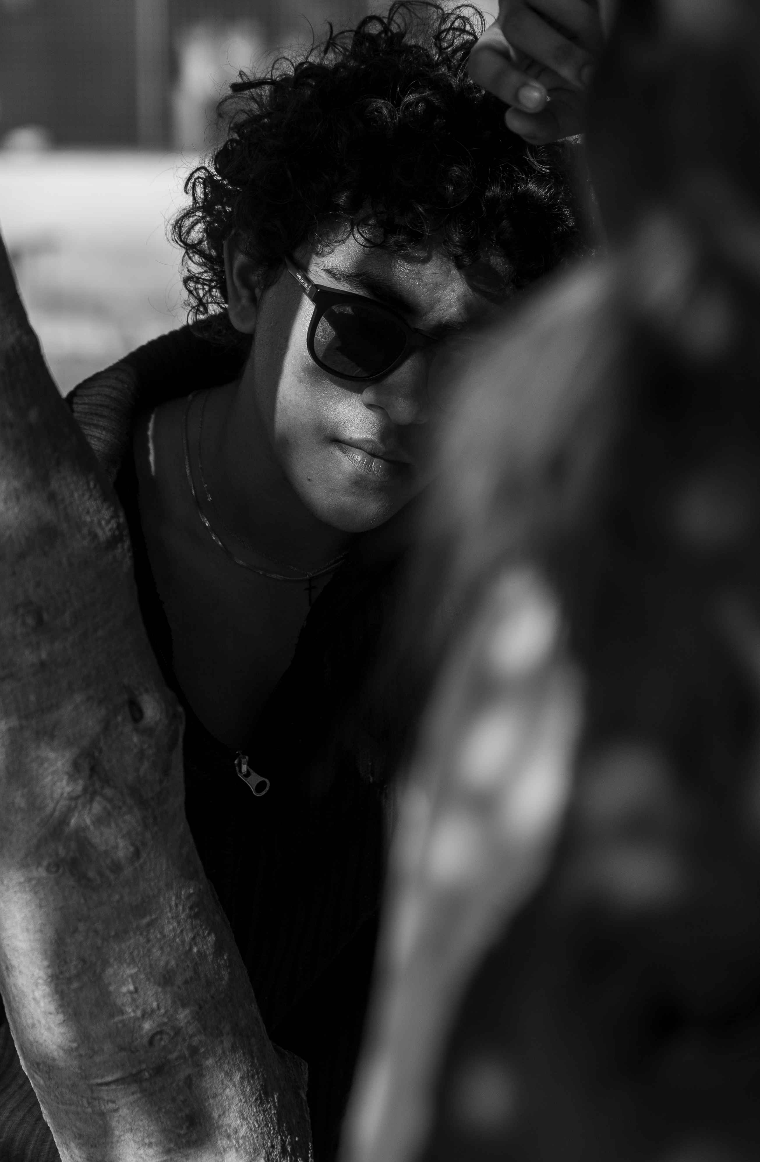 Black-and-white close portrait of a person in dark glasses beside a tree trunk.