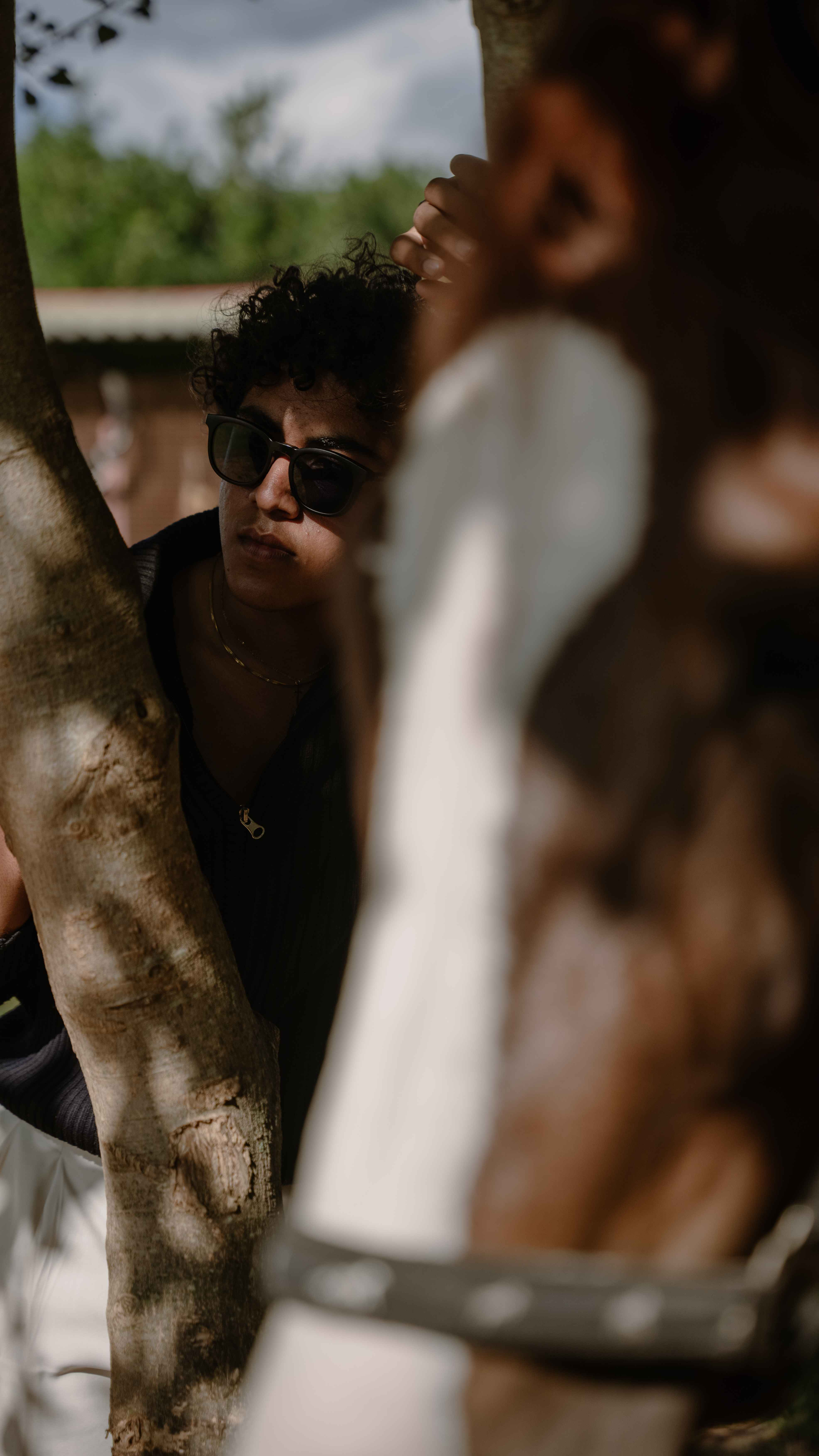 Outdoor portrait of a person in dark glasses framed by a tree trunk.