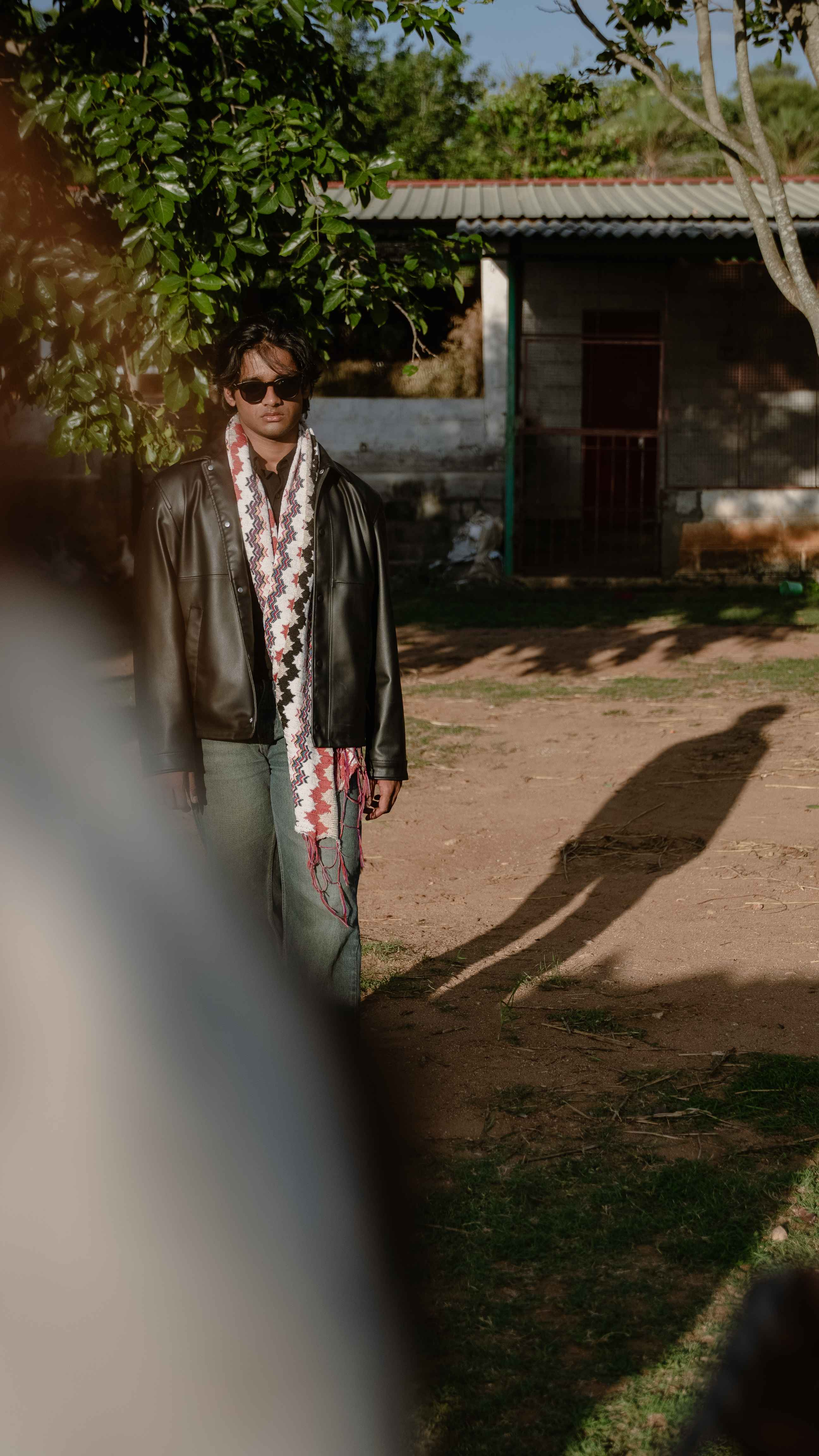 Man in sunglasses, a black jacket and patterned scarf standing outdoors near a rural farmhouse.