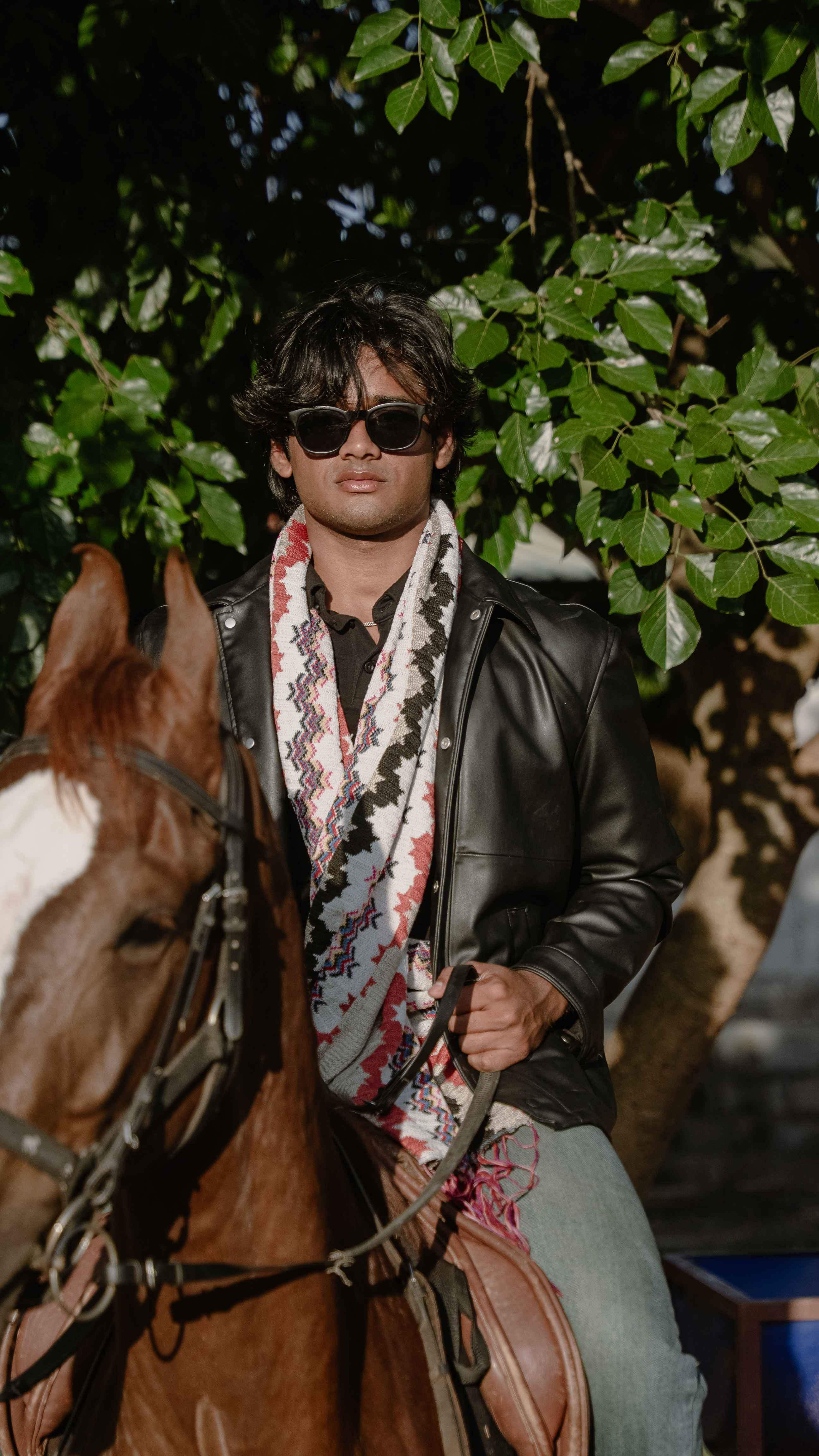 Man in sunglasses and a patterned scarf seated on a brown horse outdoors.