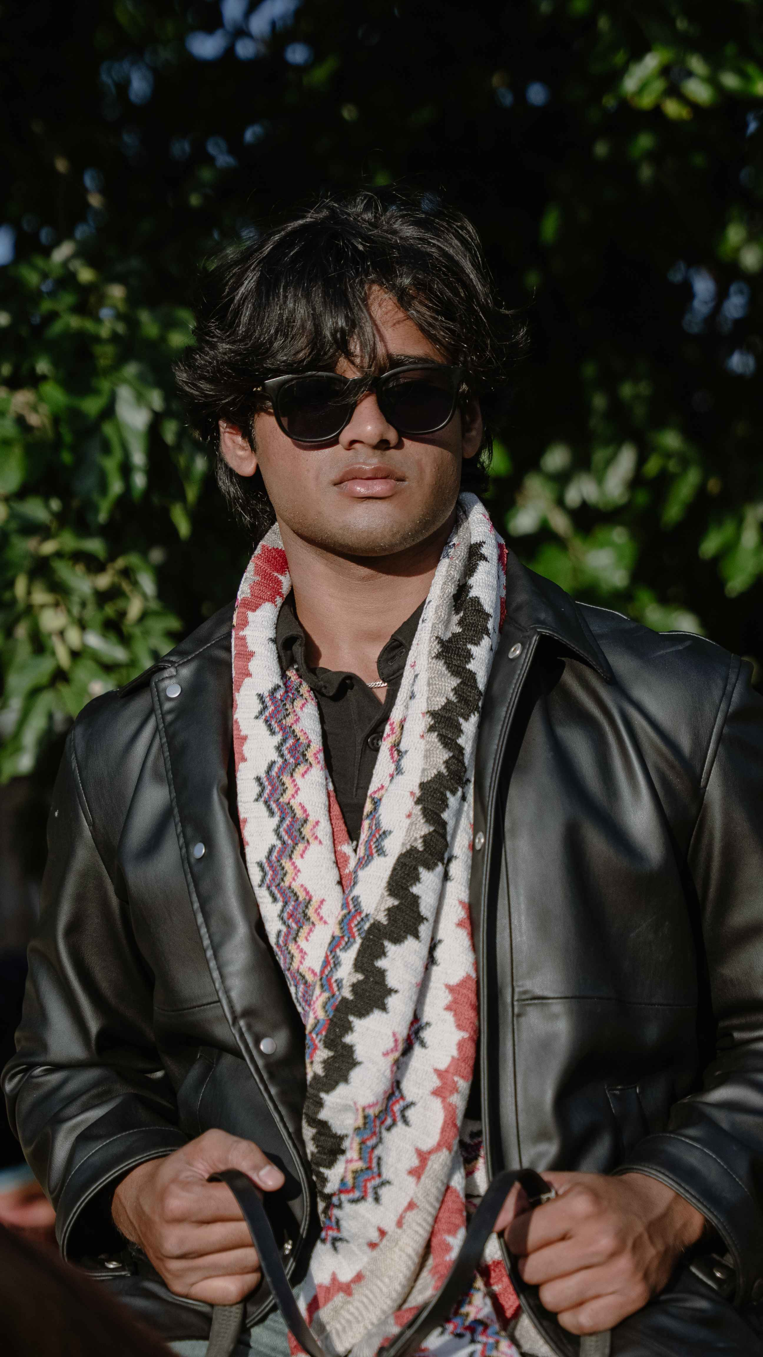 Close portrait of a man in sunglasses, a black jacket and patterned scarf against green foliage.