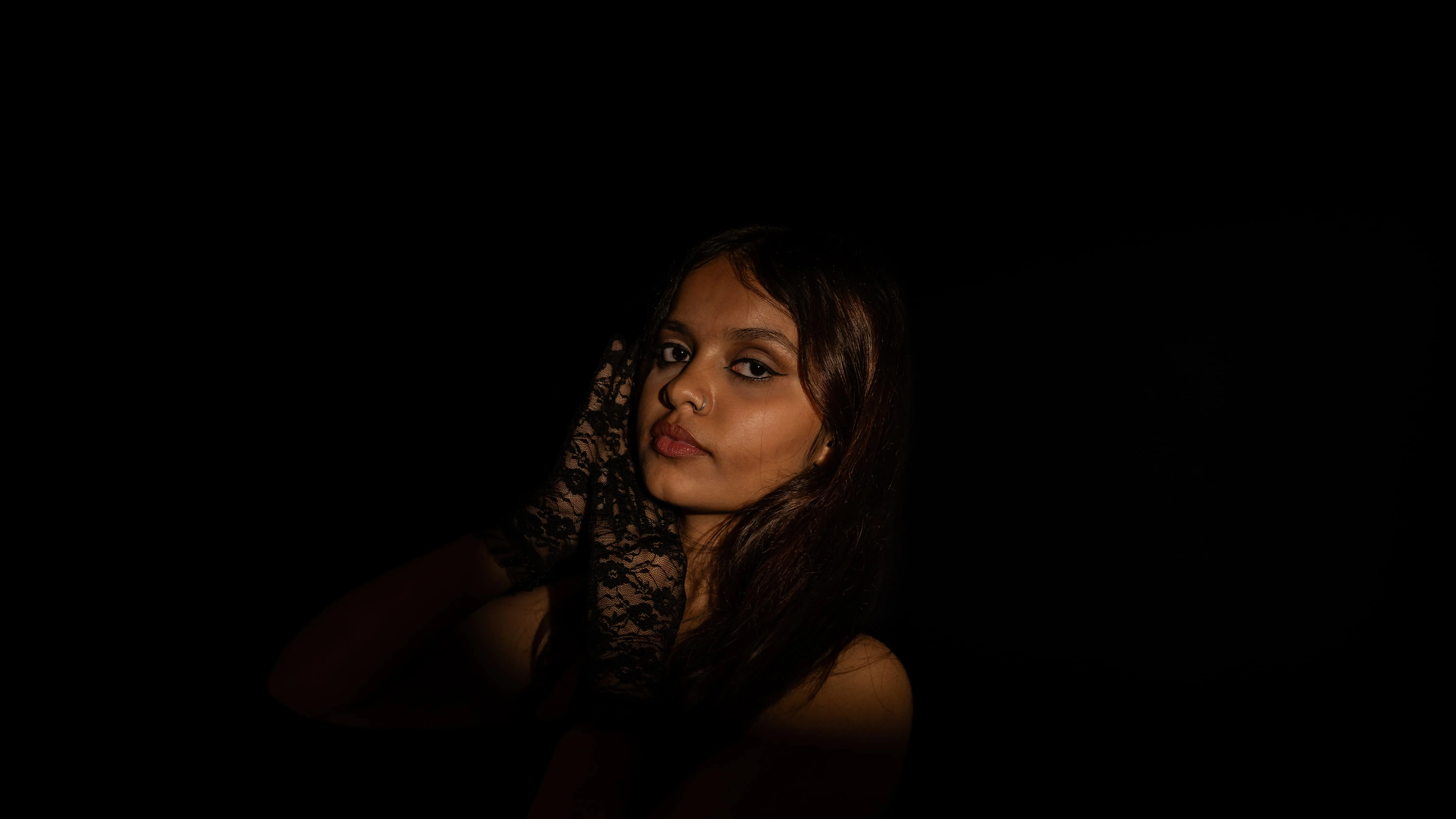Low-lit portrait of a woman in black with a lace glove against a black background.