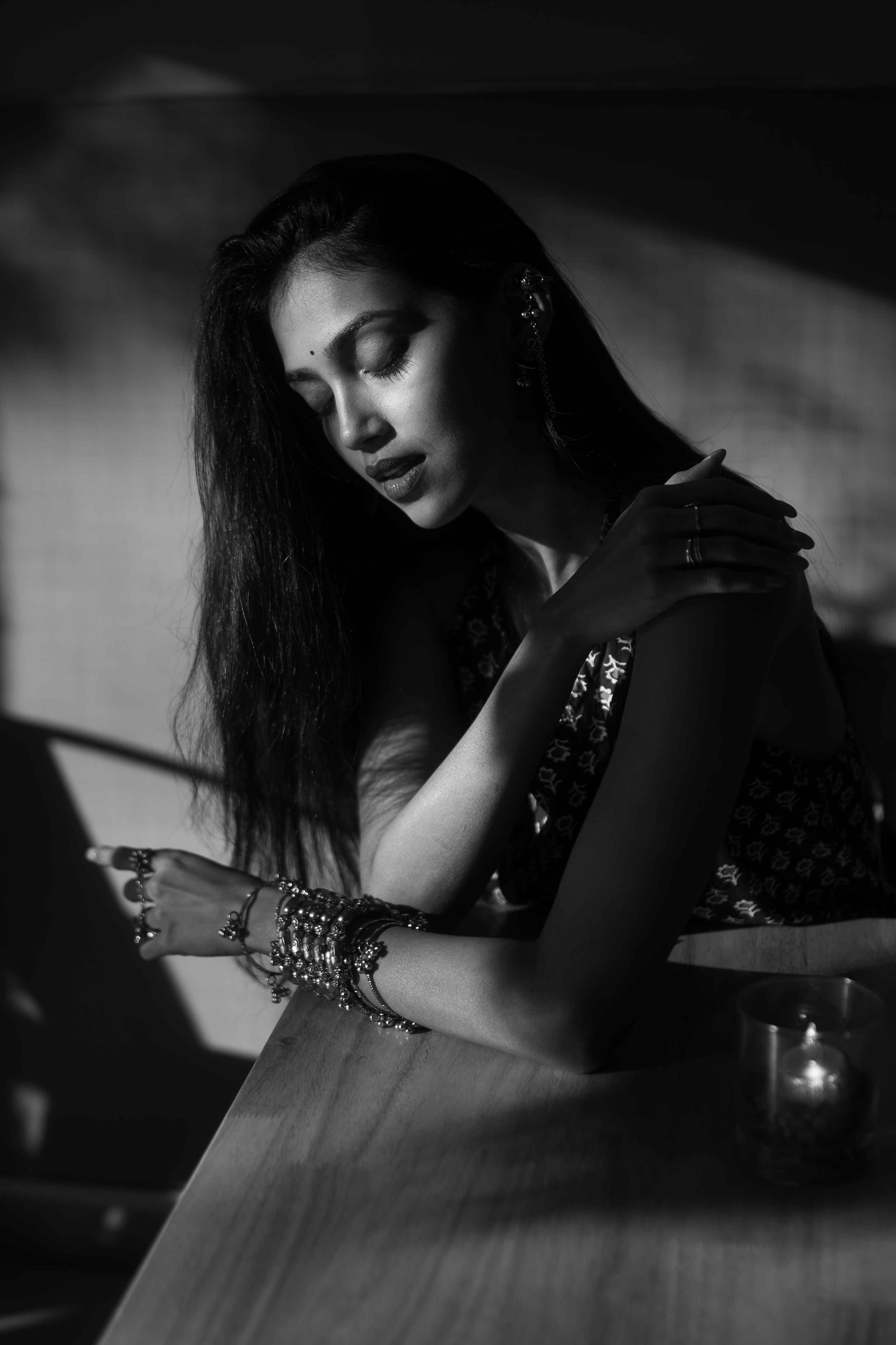 Black-and-white seated portrait of a woman wearing layered jewellery beside a table.