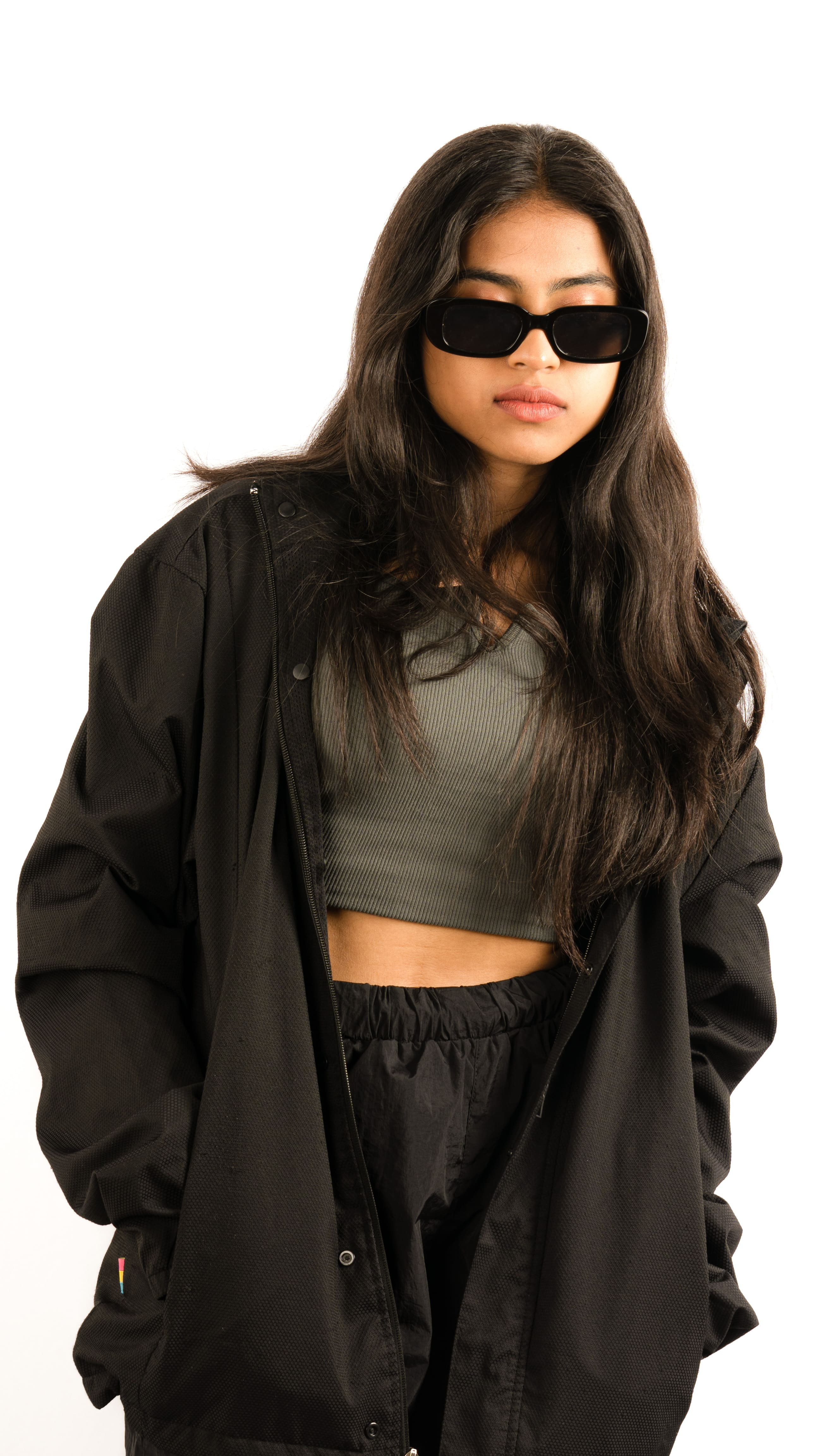 Woman in an oversized black jacket and olive crop top with sunglasses in a white studio portrait.