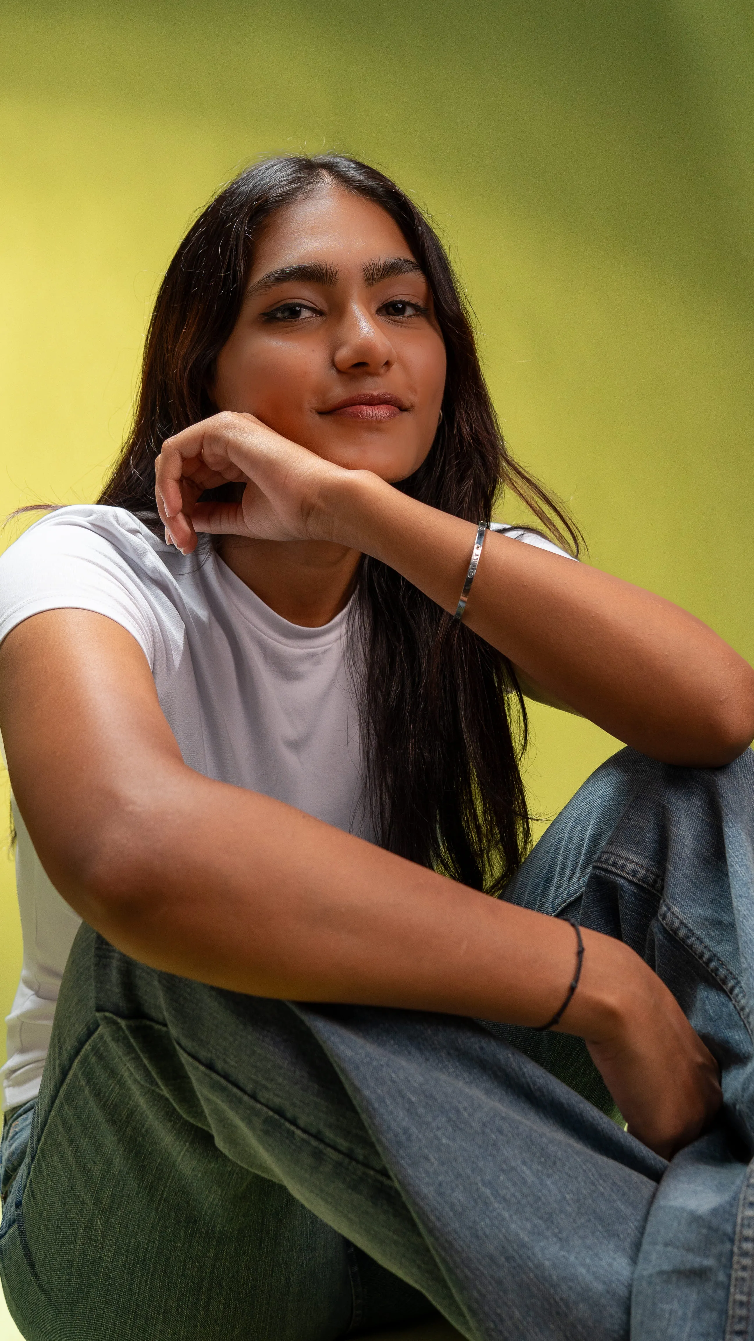 Woman in a white T-shirt and blue jeans seated against a yellow-green background.