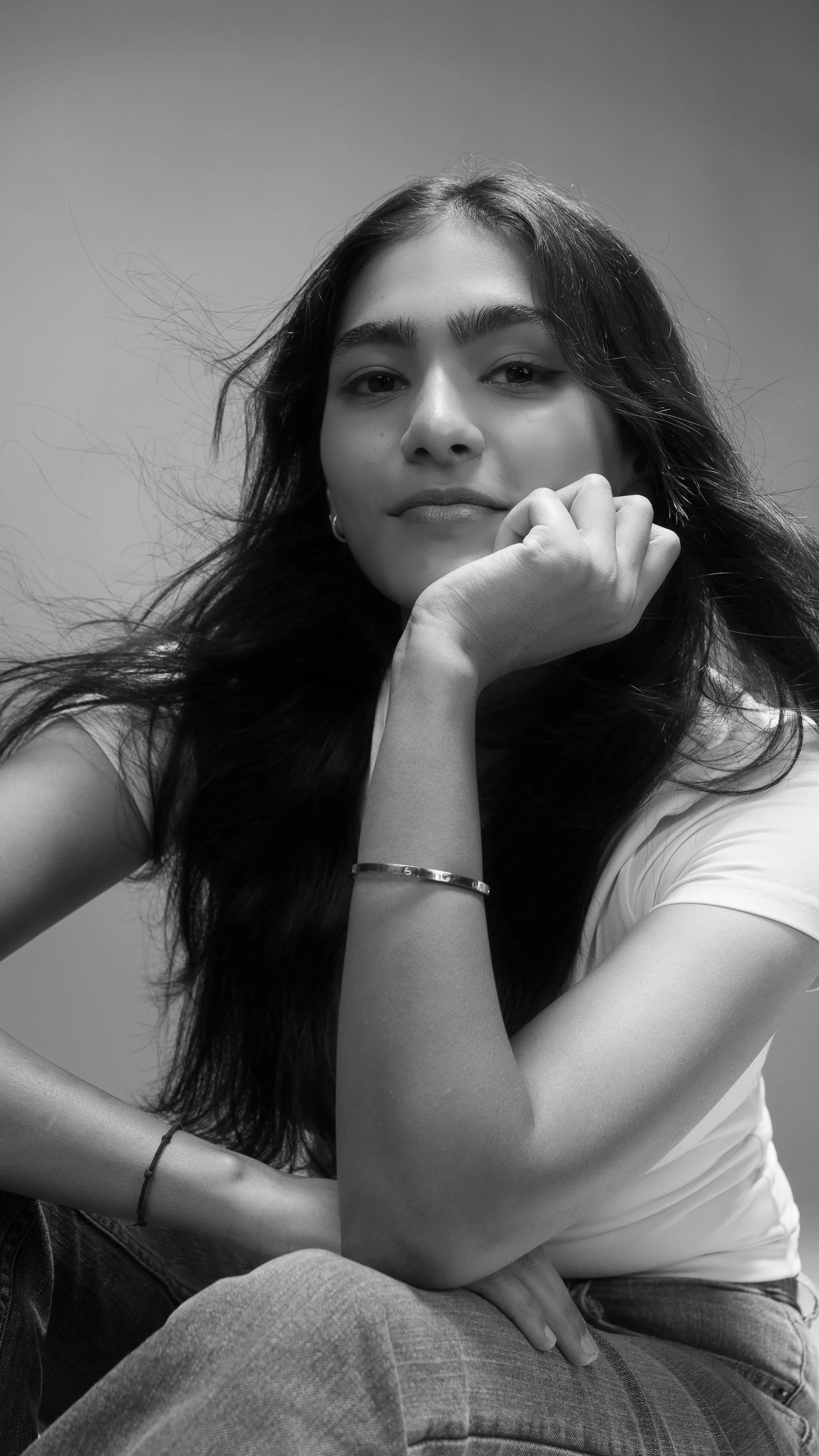 Black-and-white close portrait of a woman in a white T-shirt resting her chin on one hand.