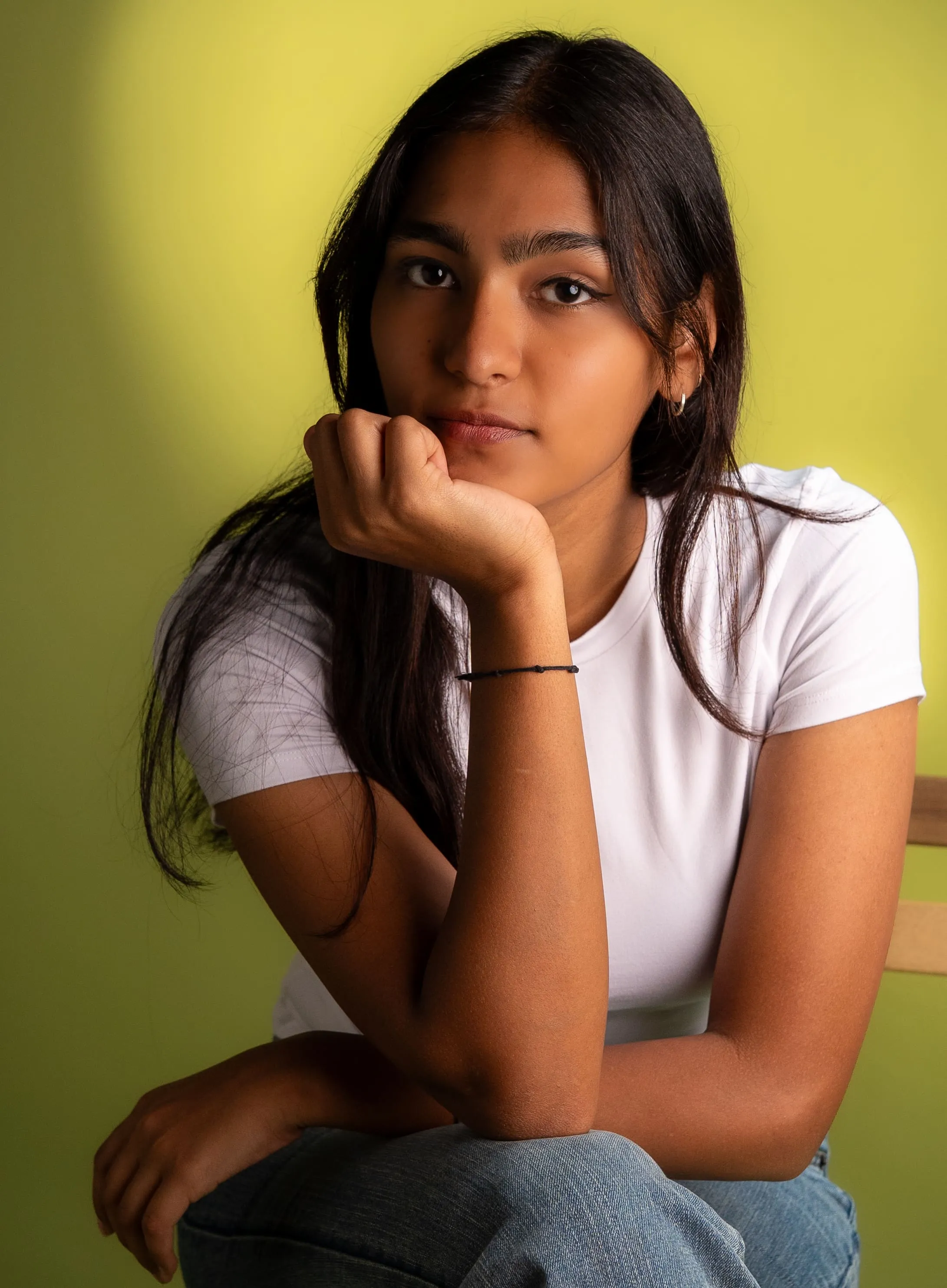 Woman in a white T-shirt and blue jeans resting her chin on one hand in a studio portrait.