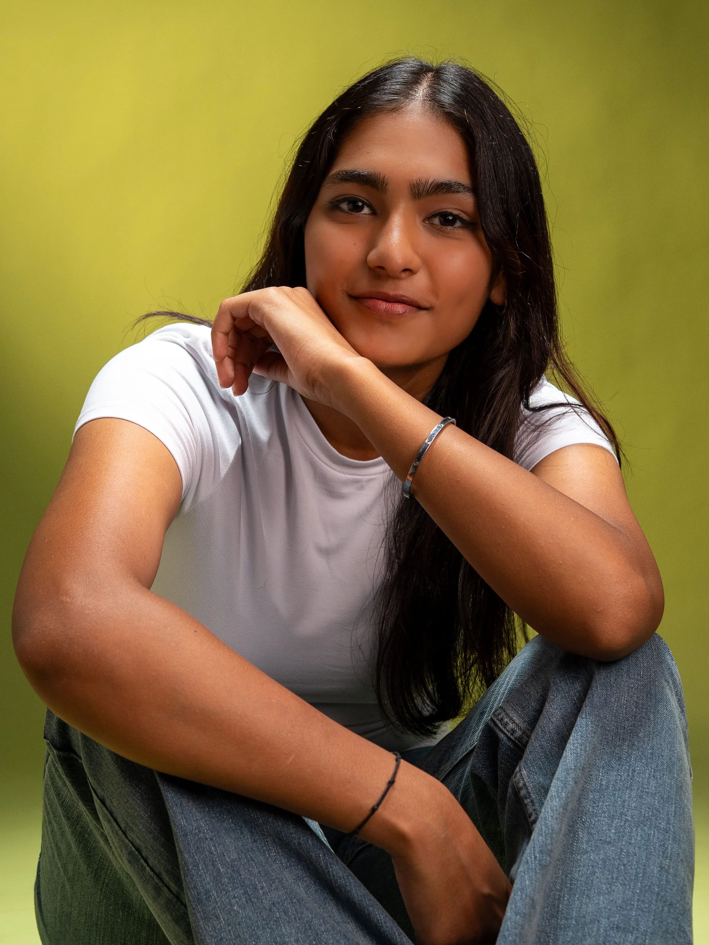 Woman in a white T-shirt and blue jeans seated against a yellow-green studio background.