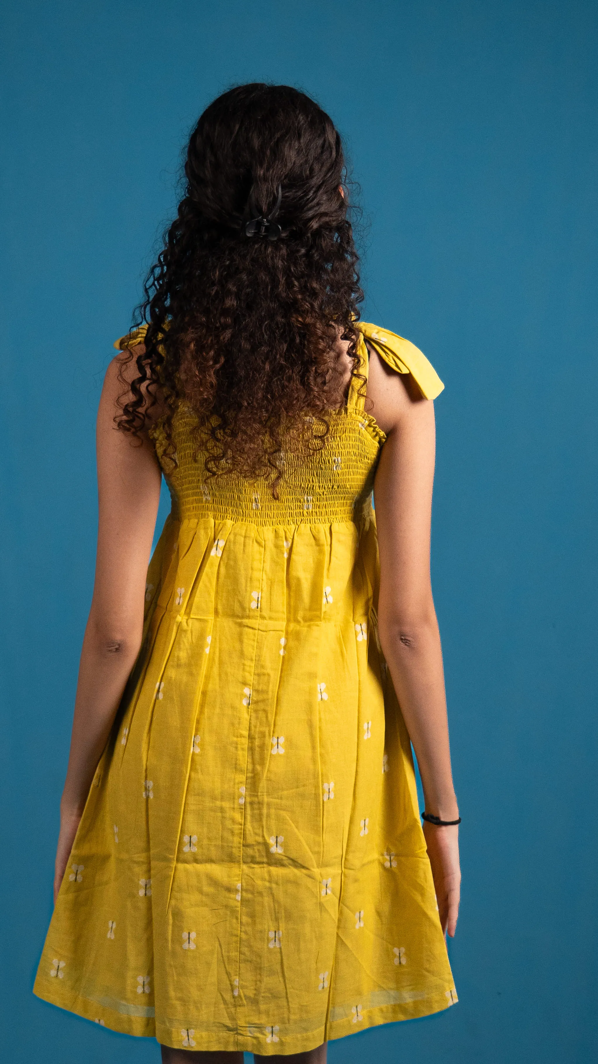 Back view of a yellow smocked dress with shoulder ties for Indyvarna.